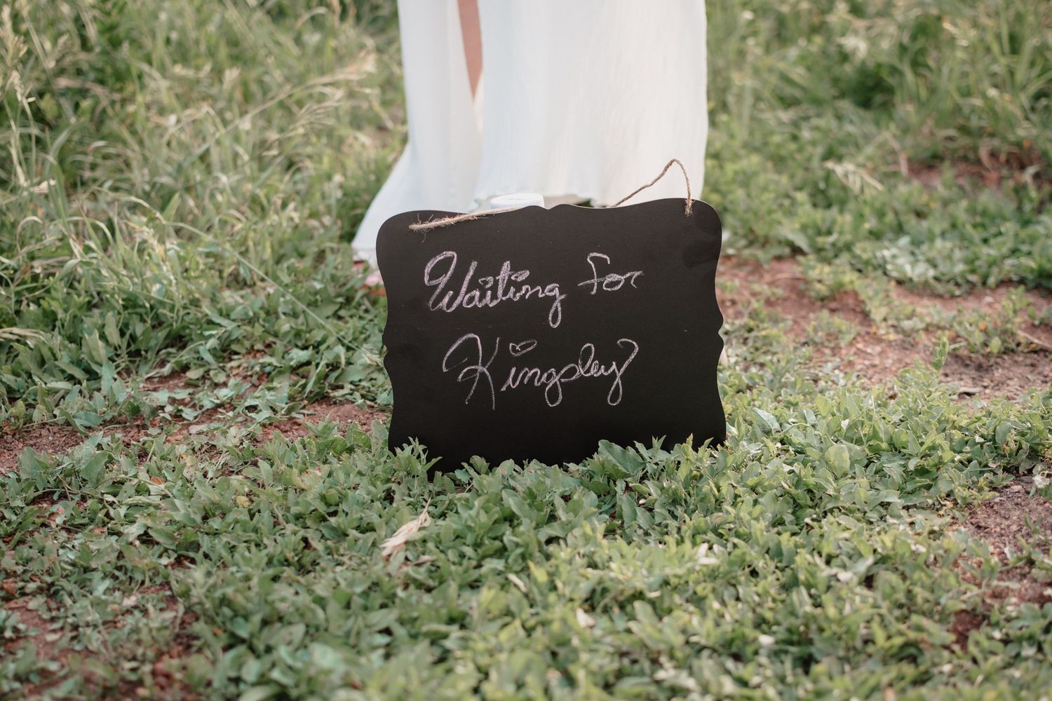 A black chalkboard wedding sign with cursive text sits on grass in an outdoor setting.