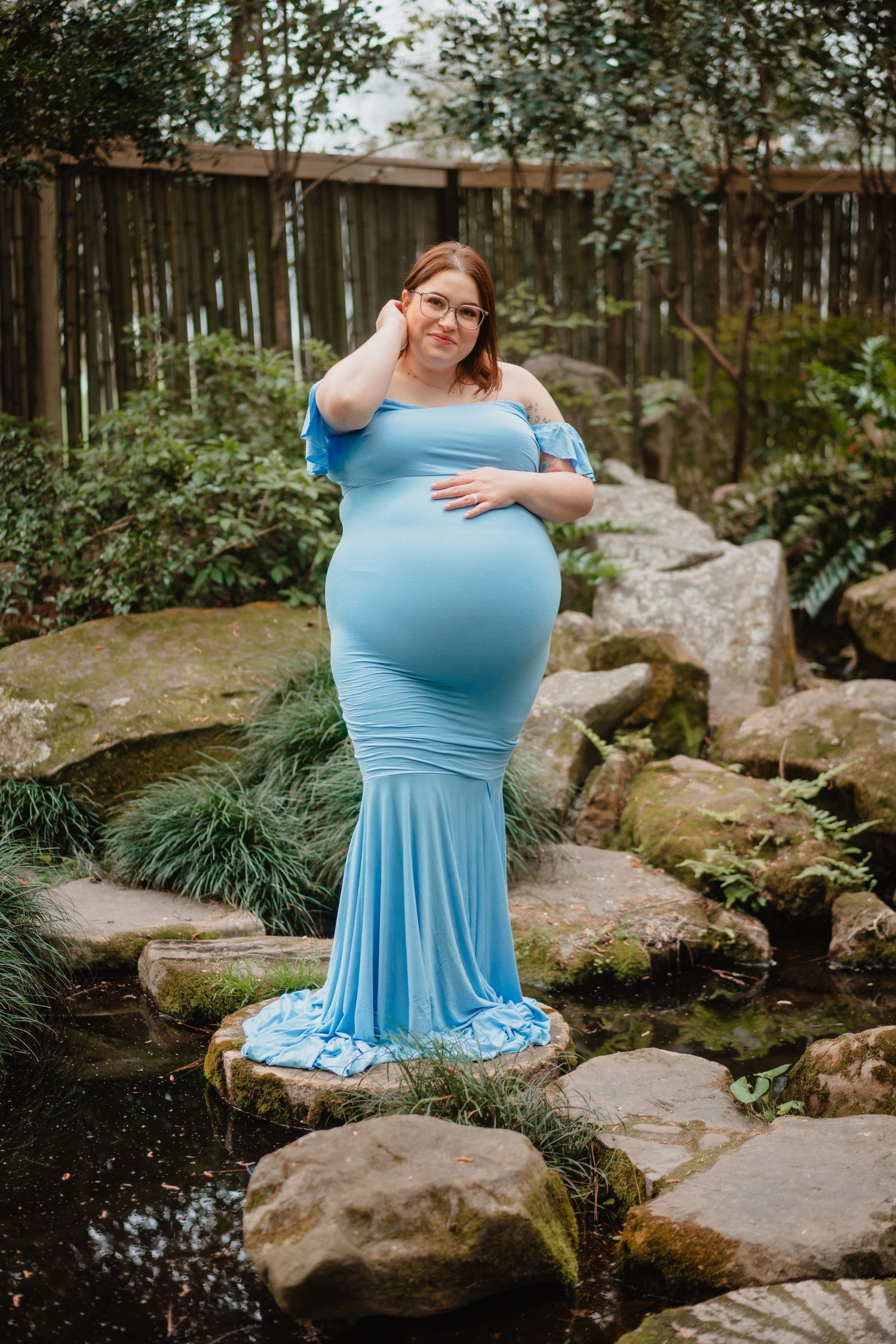 A pregnant woman wearing a blue dress stands on a path surrounded by rocks and greenery.