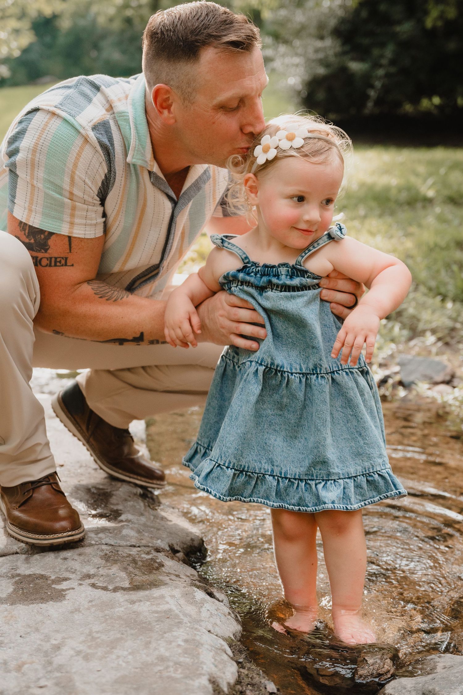 Father kissing his toddler daughter in a denim dress on rocks by a creek in a lush green park.