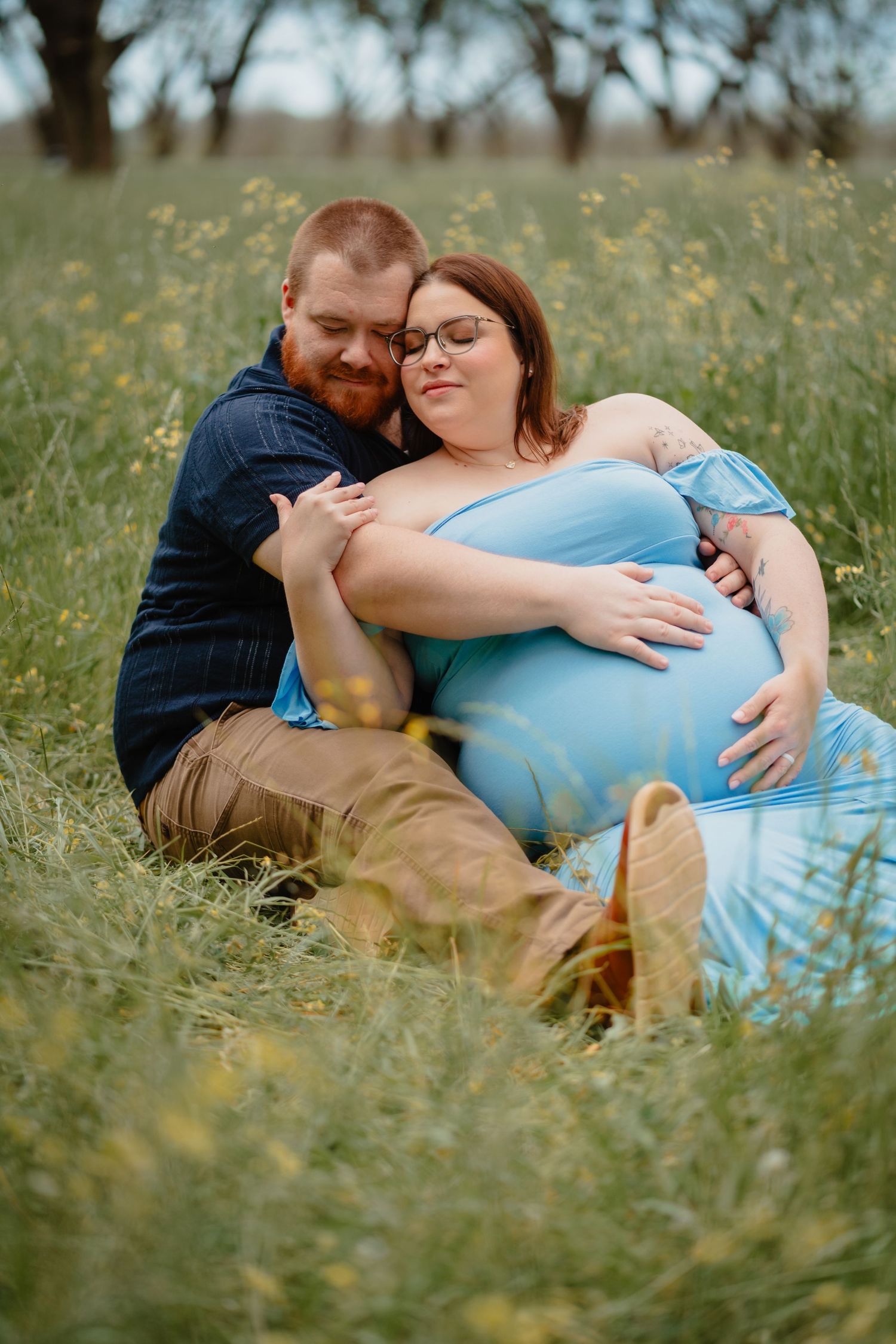 A pregnant woman is embraced by her partner in a grassy field surrounded by trees.