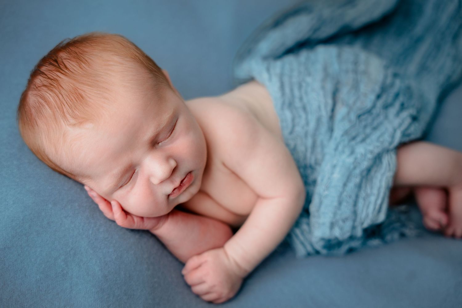 Peaceful newborn baby with red hair sleeping on a soft blue blanket, wrapped in a light blue swaddle cloth.
