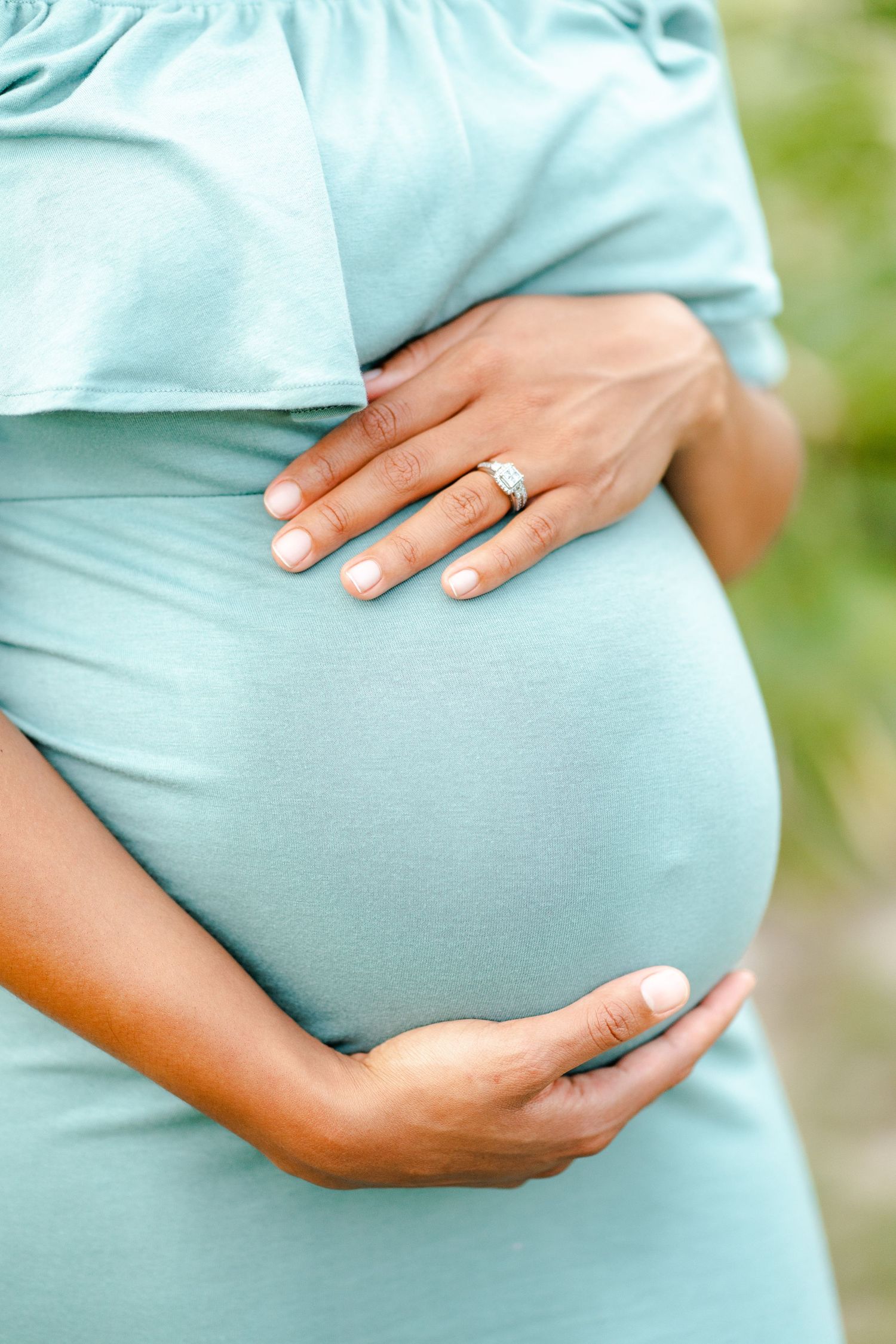 Close up of pregnant belly in mint green dress with hands cradling bump and engagement ring visible.