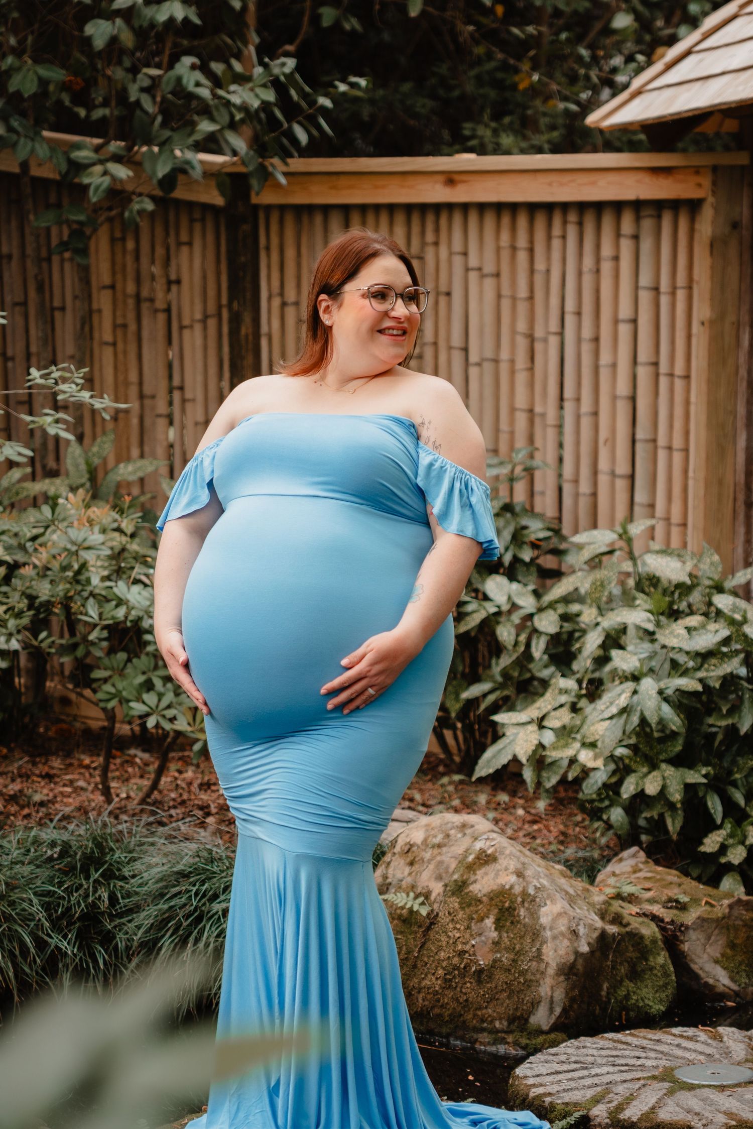 A pregnant woman wearing a blue dress stands in a garden surrounded by greenery.