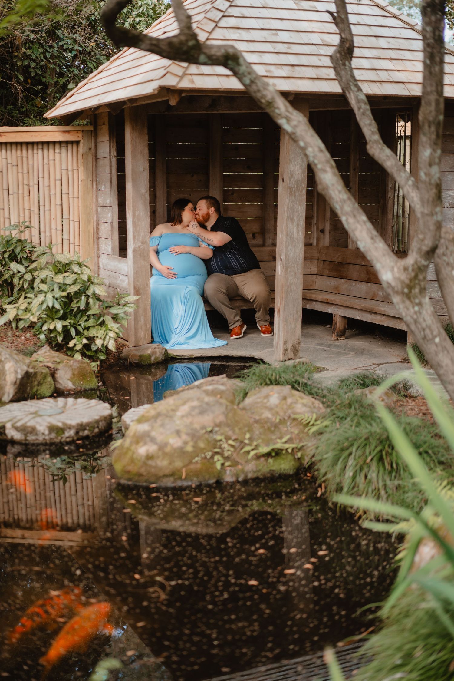 A couple embracing on the porch of a wooden cabin surrounded by lush greenery and a koi pond.