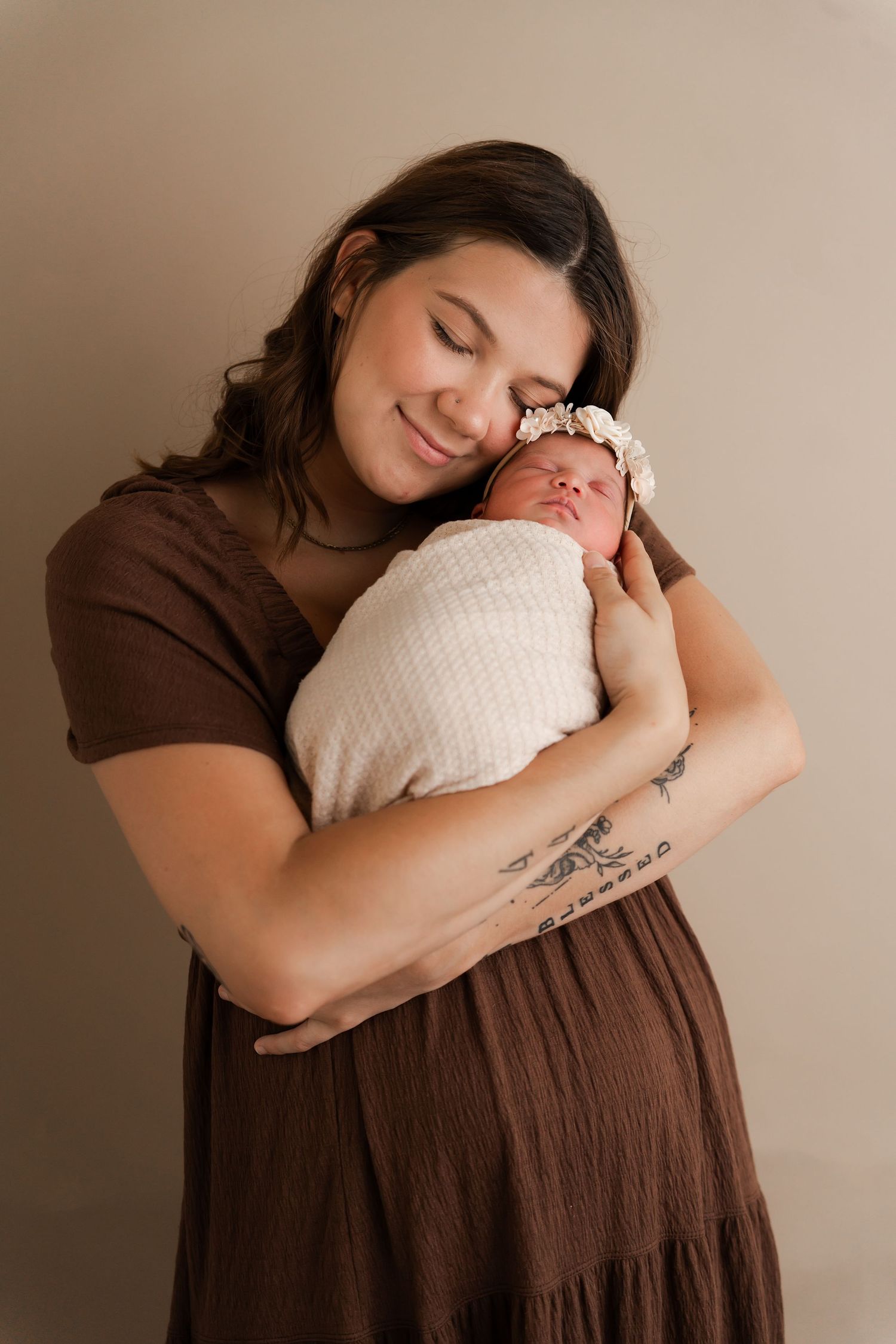 A smiling mother in a brown dress tenderly holds her swaddled newborn baby girl wearing a floral headband.