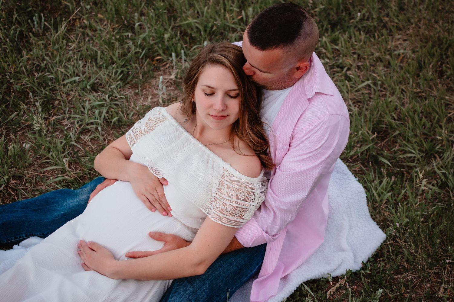 Couple in romantic outdoor engagement photos with bride in white dress and groom in pink shirt laying in grass.