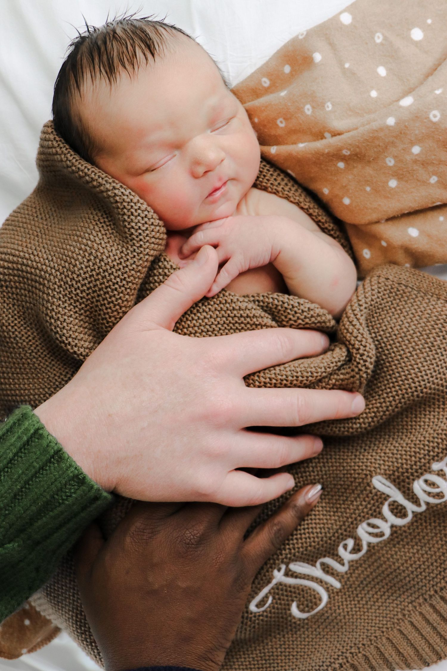 A sleeping newborn baby swaddled in a brown knit blanket with polka dot fabric and gentle hands holding them.