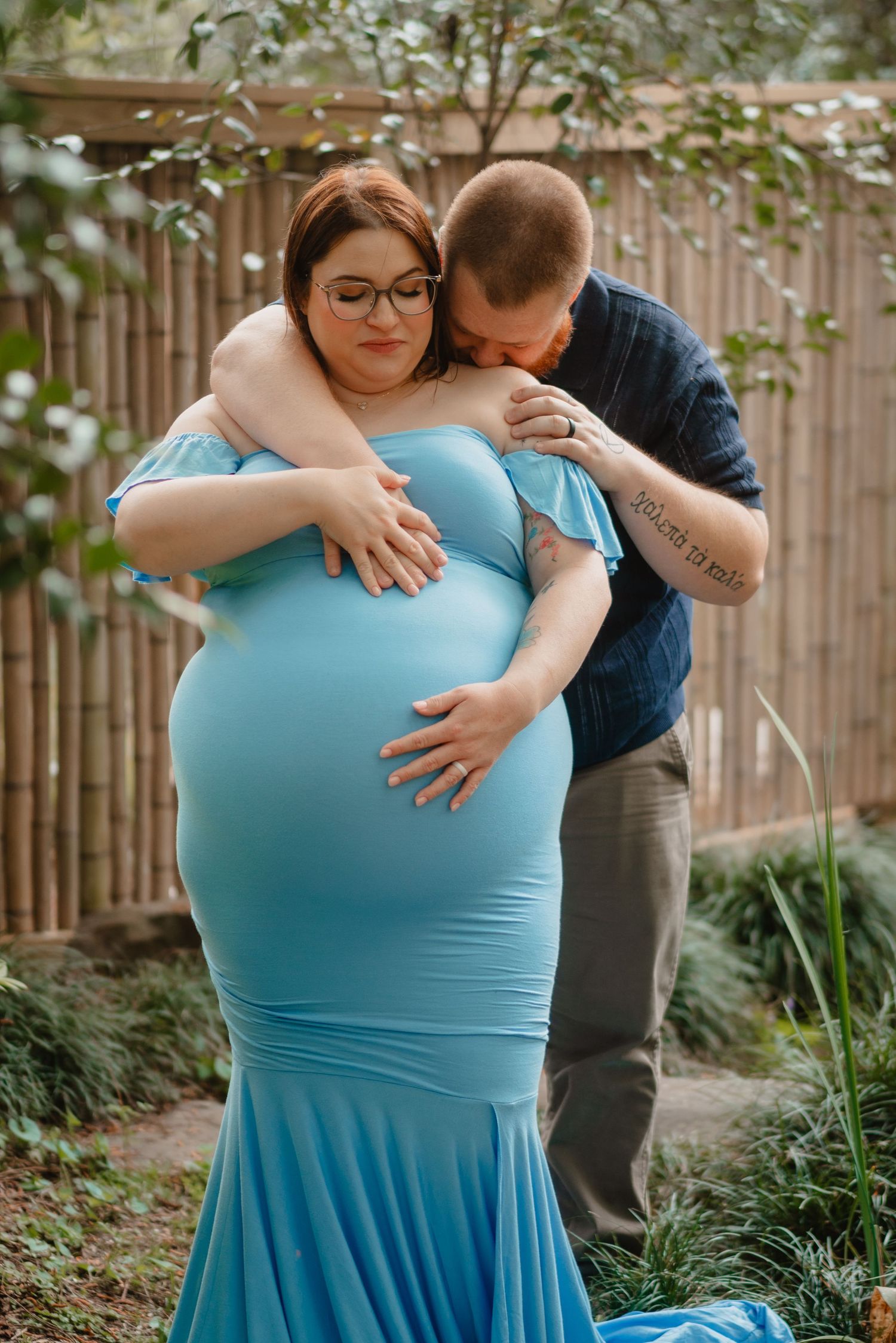 A pregnant woman wearing a blue dress is embraced by her partner in a garden setting.