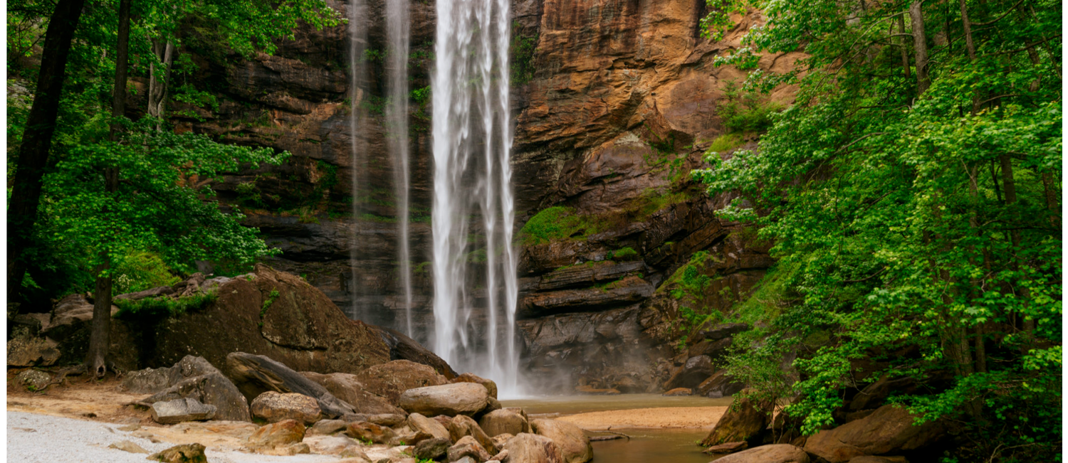 A stunning waterfall cascades down red rock cliffs surrounded by lush green trees and boulders in a serene forest setting.