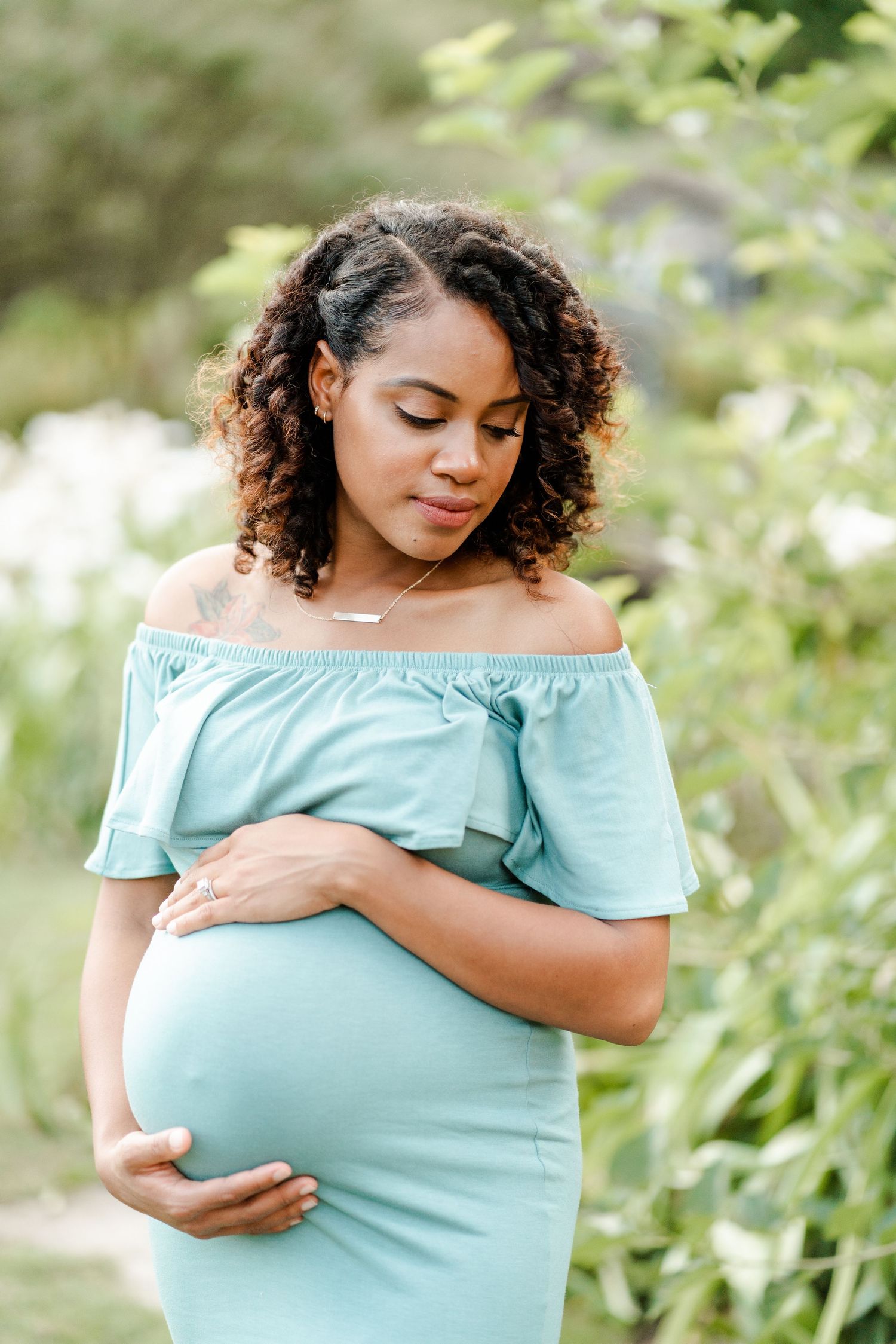 Expectant mother in light blue off-shoulder dress cradles baby bump while standing in garden setting.