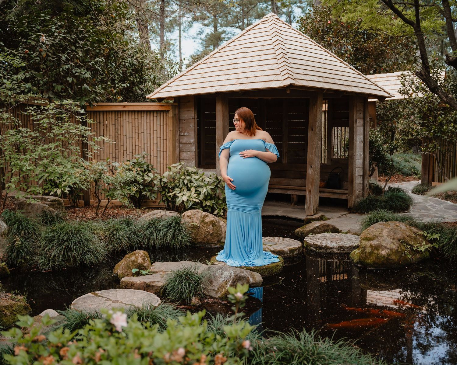 A pregnant woman wearing a blue dress stands in a scenic garden with a wooden gazebo in the background.