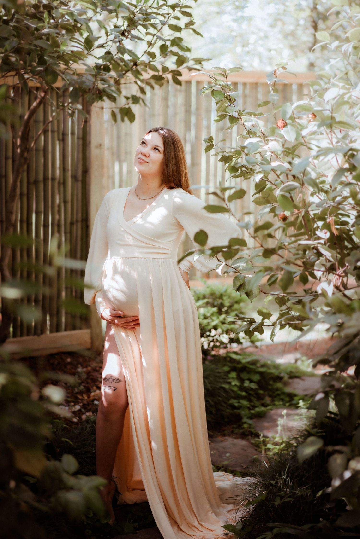 Pregnant woman in elegant white dress standing in a sunlit garden surrounded by lush green plants and blooming roses.