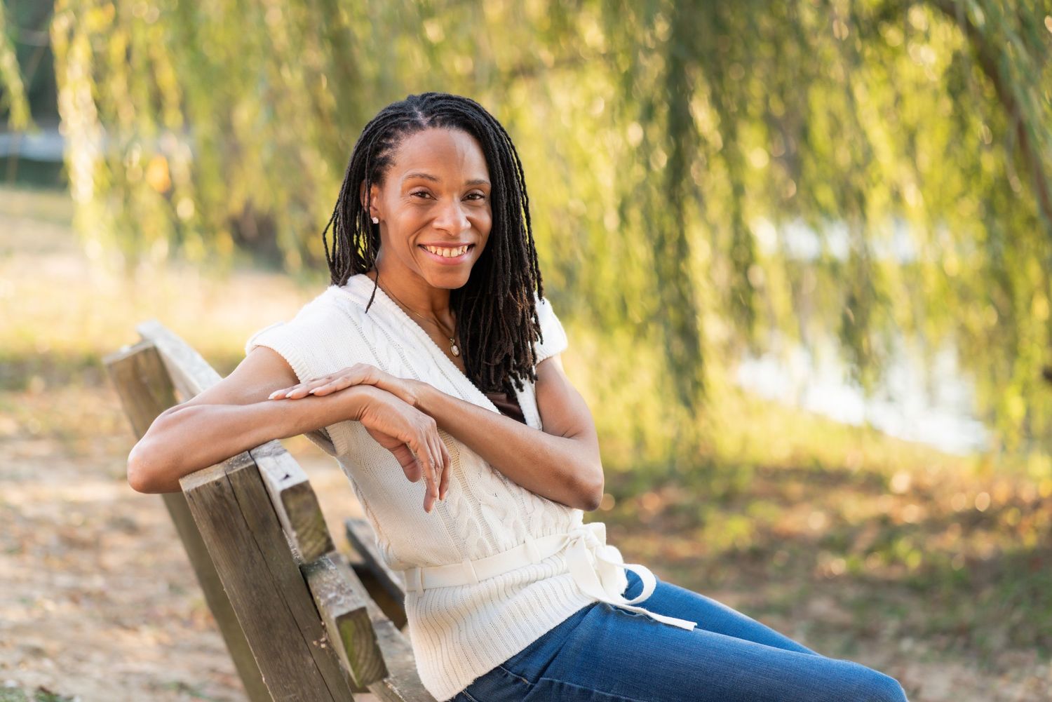 Person relaxing on a wooden bench in a park with willow trees in the background.