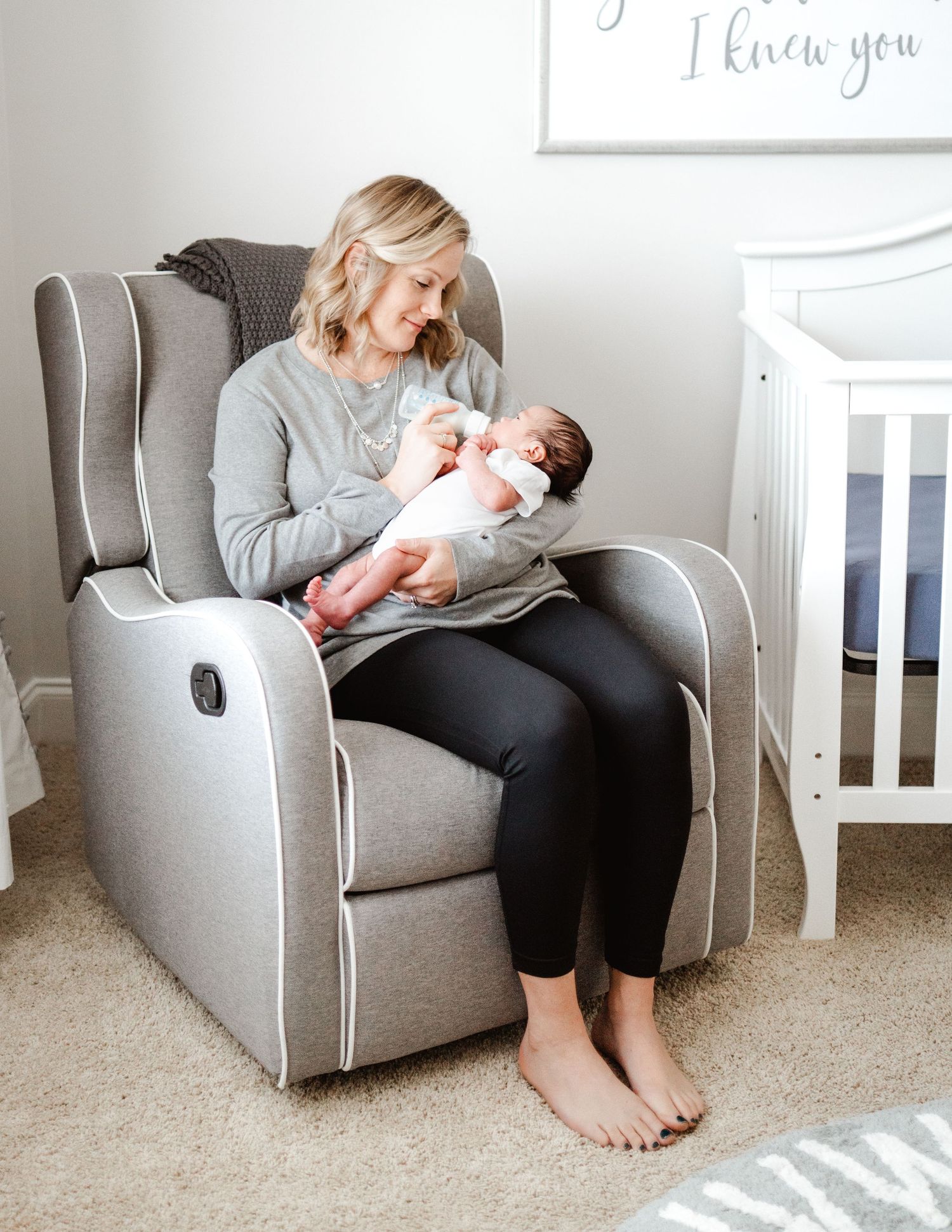 Parent sitting in gray nursery recliner while holding sleeping newborn.