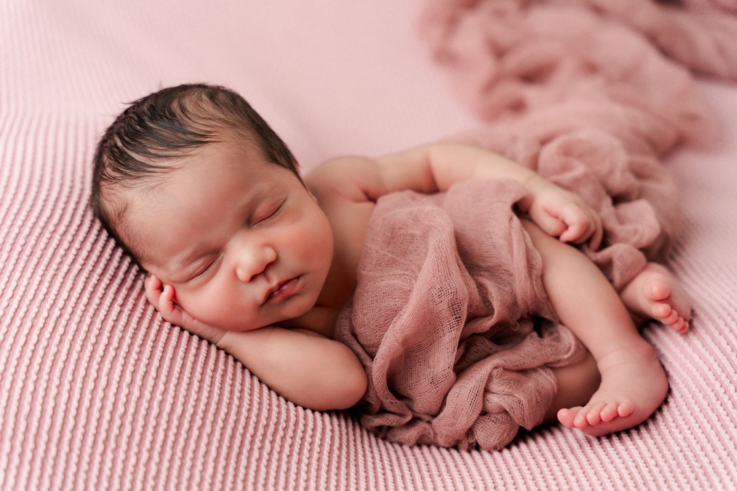 Peaceful newborn baby sleeping curled up on a soft pink blanket, wrapped in a delicate pink gauze cloth.