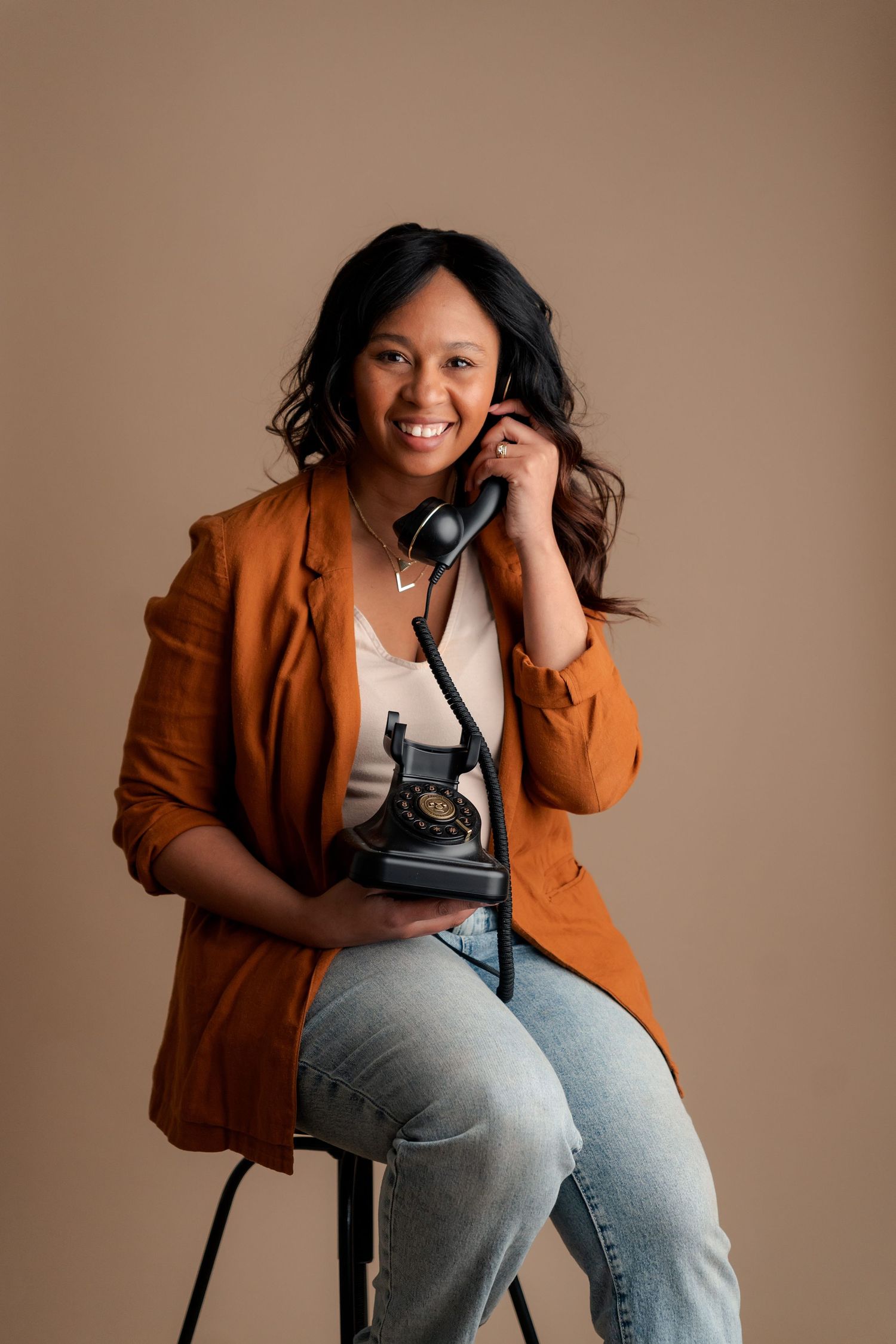 Person in brown blazer and jeans sits casually with vintage telephone against neutral backdrop.