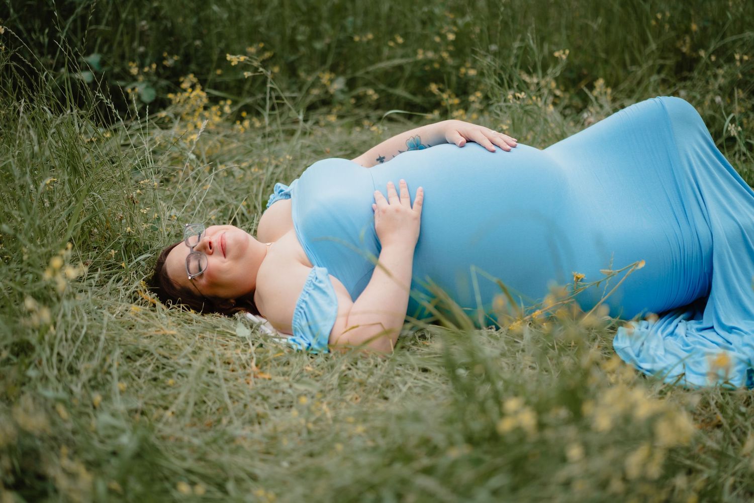 A pregnant woman in a blue dress resting in a grassy field.