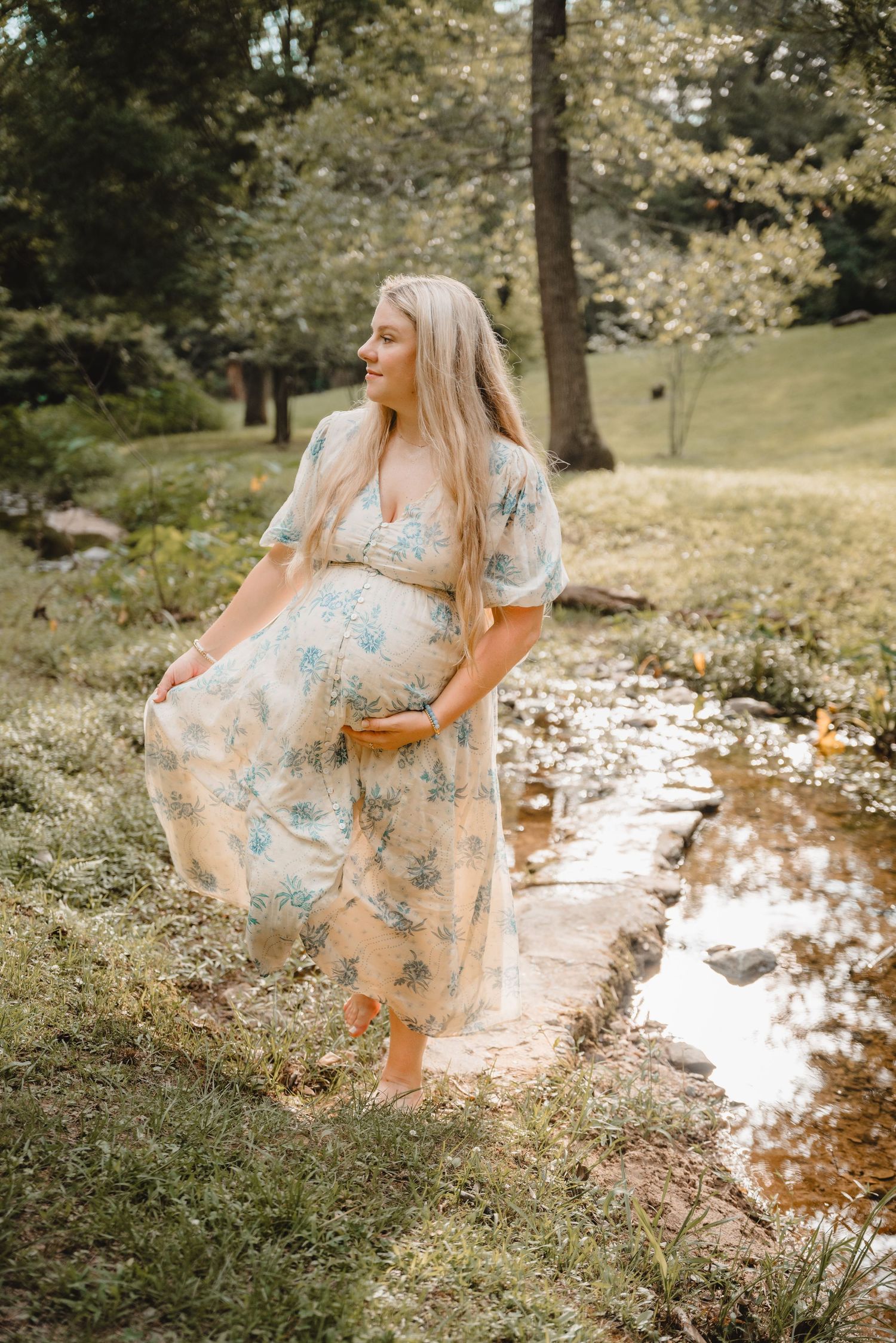 Pregnant woman in floral maxi dress standing by a stream in a lush green park.