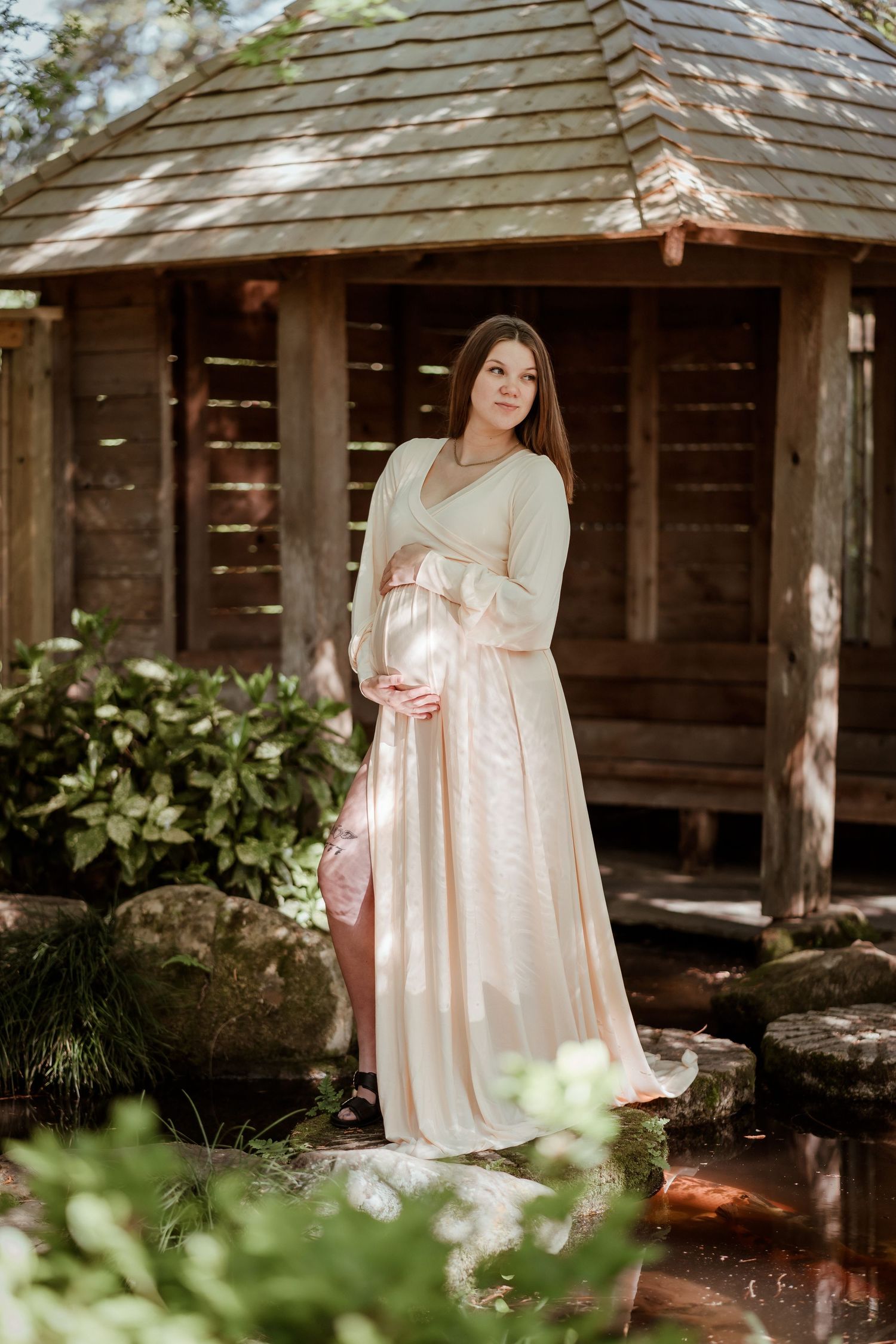 Pregnant woman in elegant blush gown cradling baby bump outdoors near wooden gazebo surrounded by lush greenery.