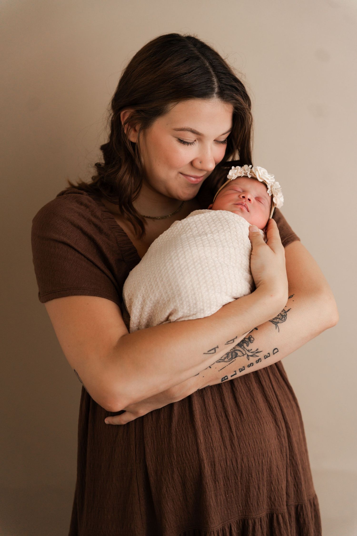 A mother in a brown dress tenderly holds her swaddled newborn with a floral headband in a studio portrait.