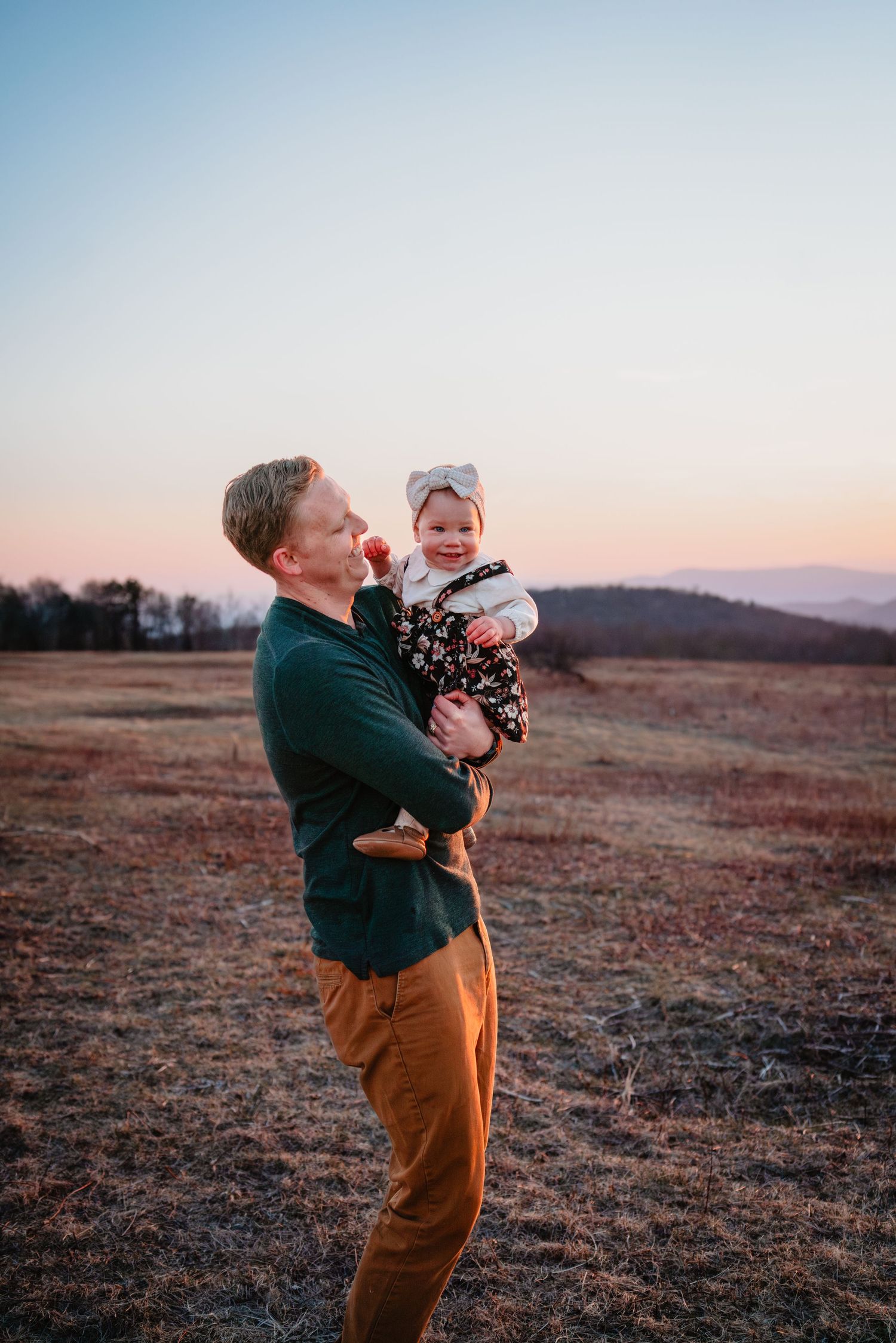 A father holds his smiling baby girl at sunset in an open field with mountain views in the background.