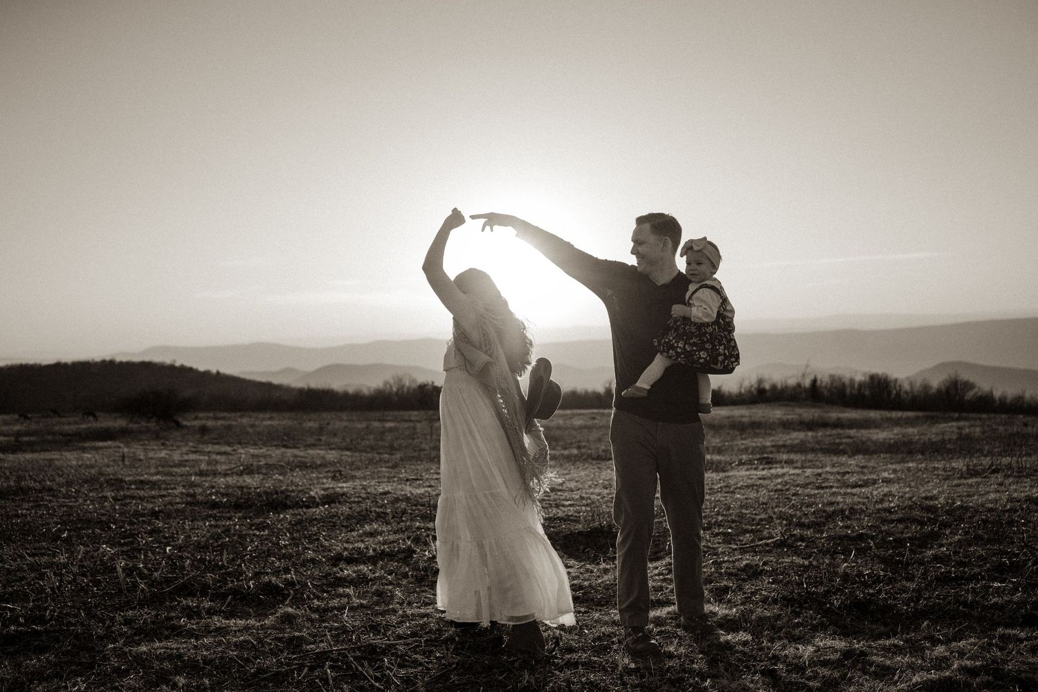 Black and white photo of a family with two children posing at sunset in an open field with rolling hills.