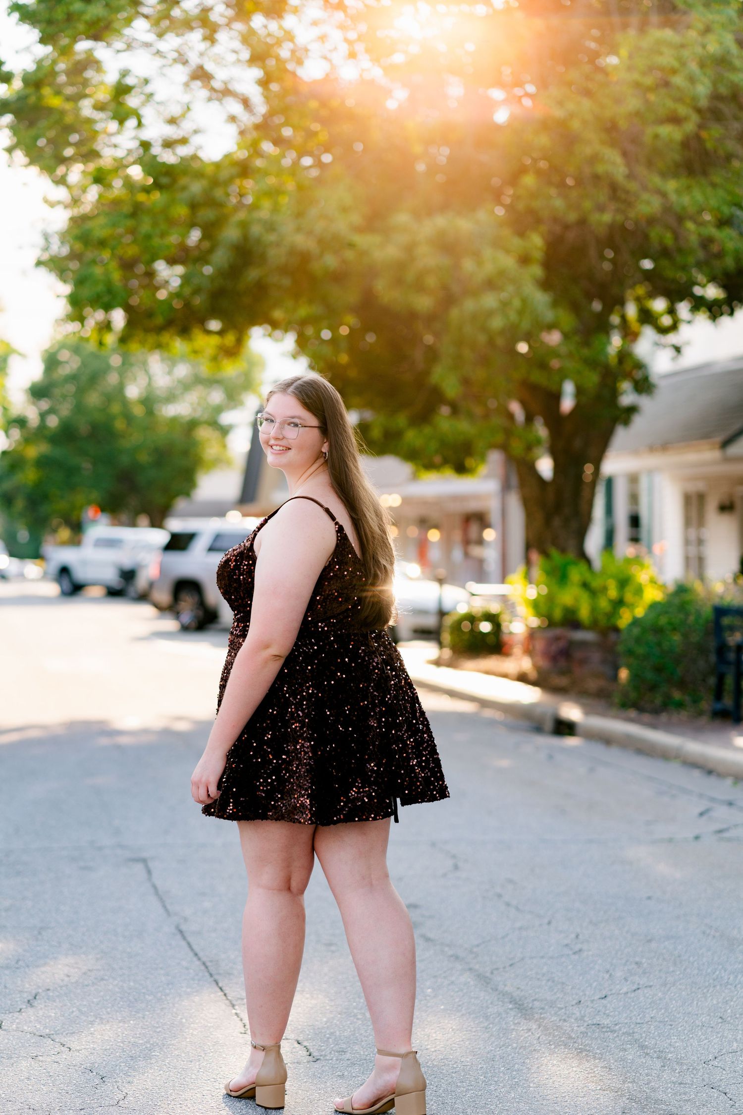 A person in a sparkly black dress poses on a sunny small town street with trees and storefronts in the background.