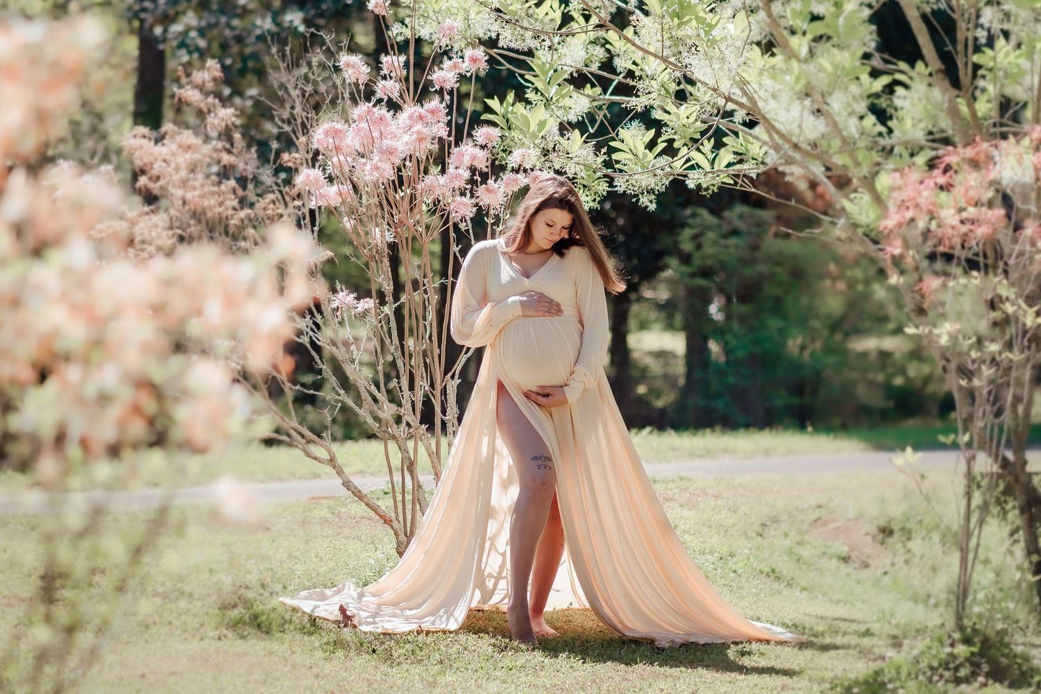 Pregnant woman in flowing peach gown cradling belly among blooming spring trees in a serene outdoor maternity photo.