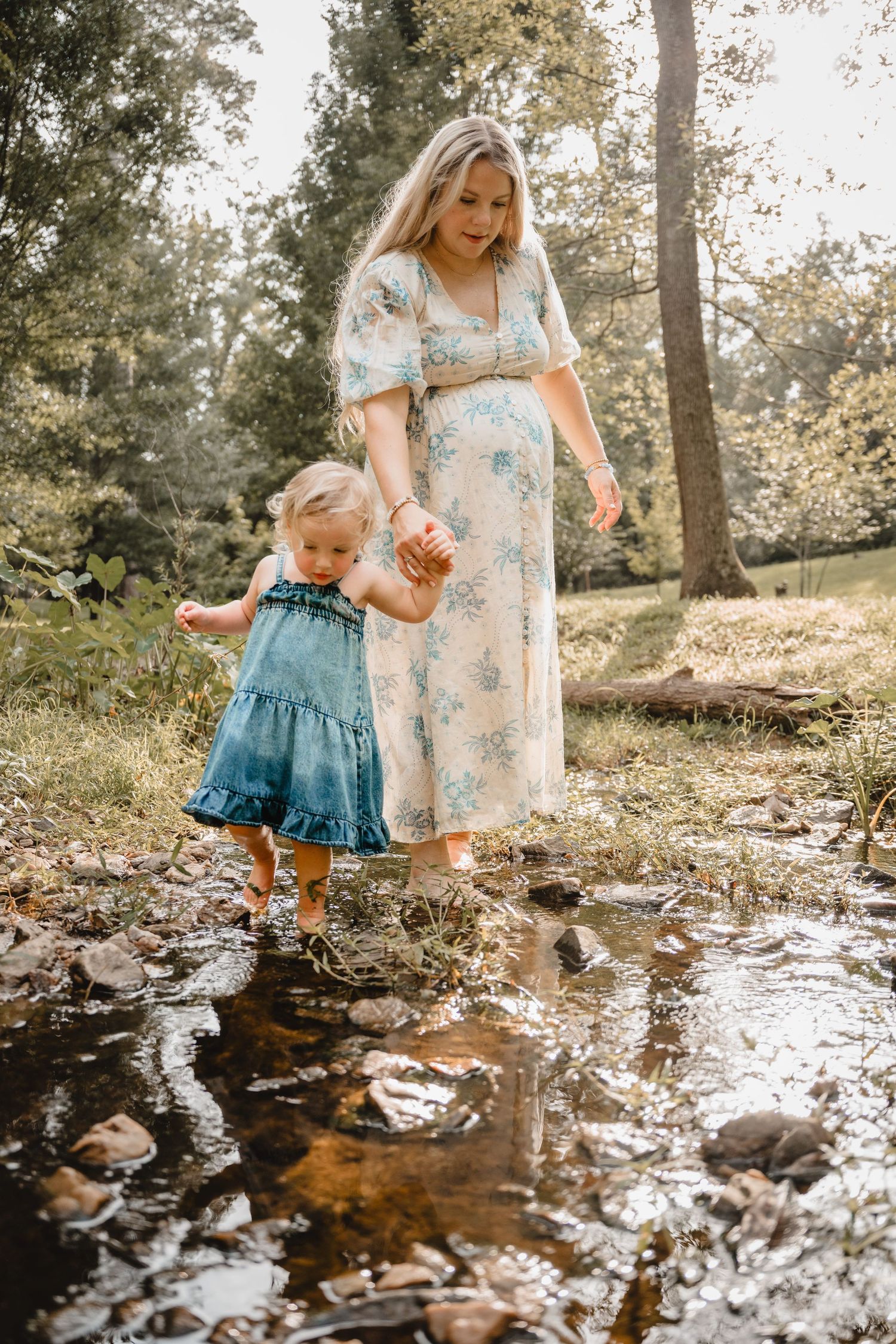 Pregnant woman in floral dress helps toddler girl wade through a shallow creek in a lush green forest.