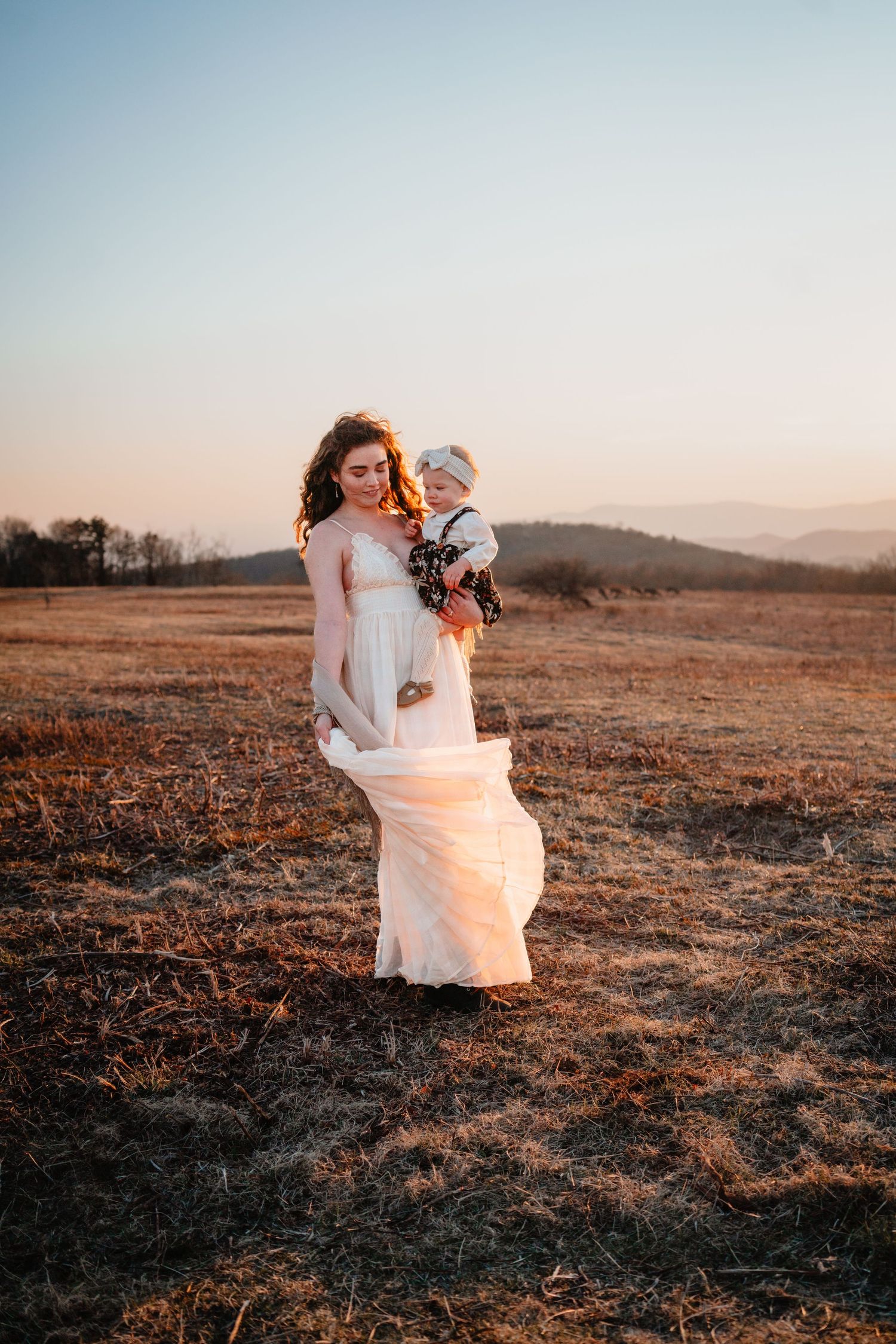 A woman in a white dress holds a baby in a golden field at sunset with mountains in the background.