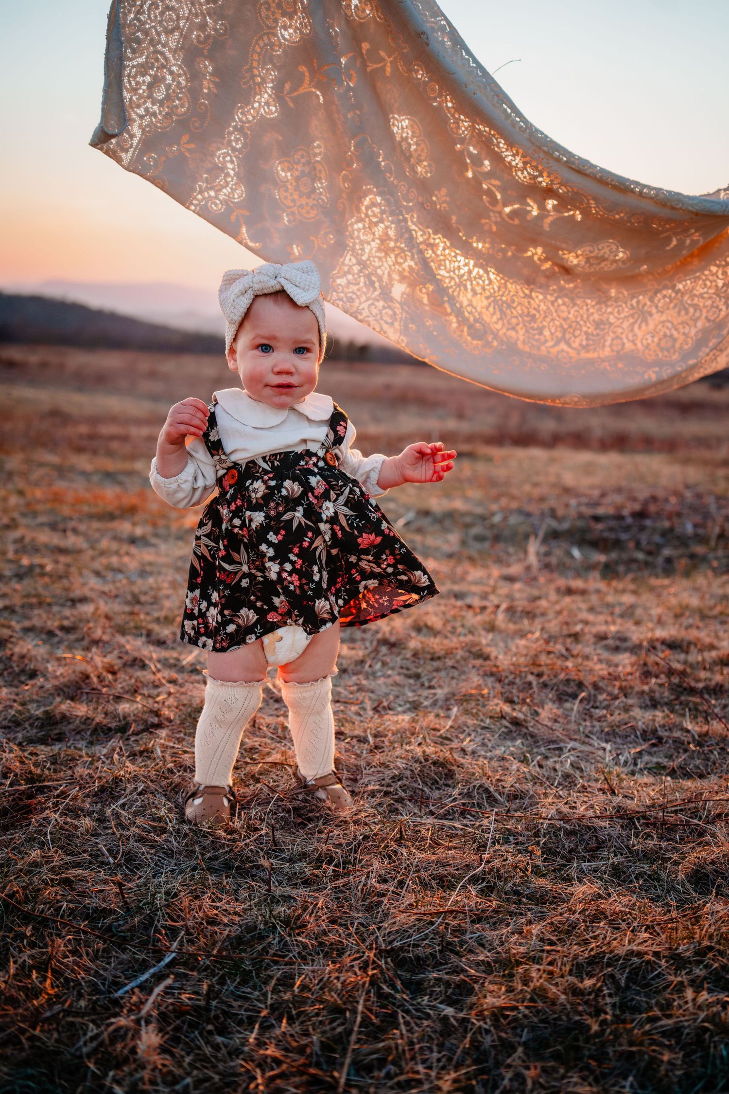 Baby girl in floral dress and bow stands in golden field at sunset with lace fabric flowing above.