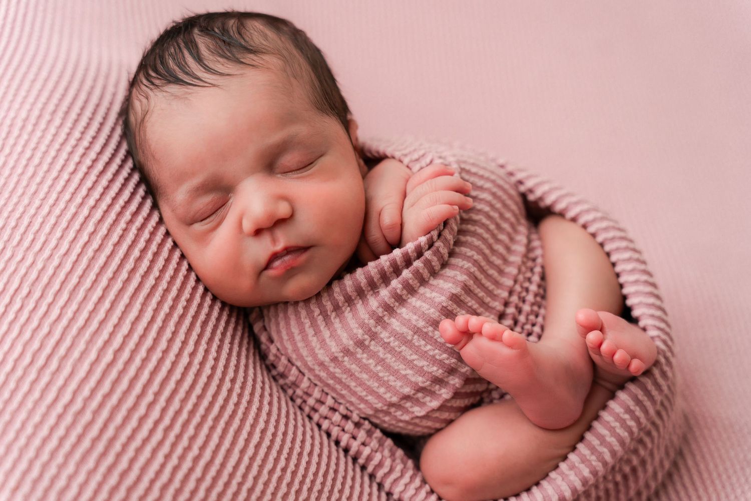 Peaceful newborn baby swaddled in a soft pink knit wrap, sleeping on a pink blanket.
