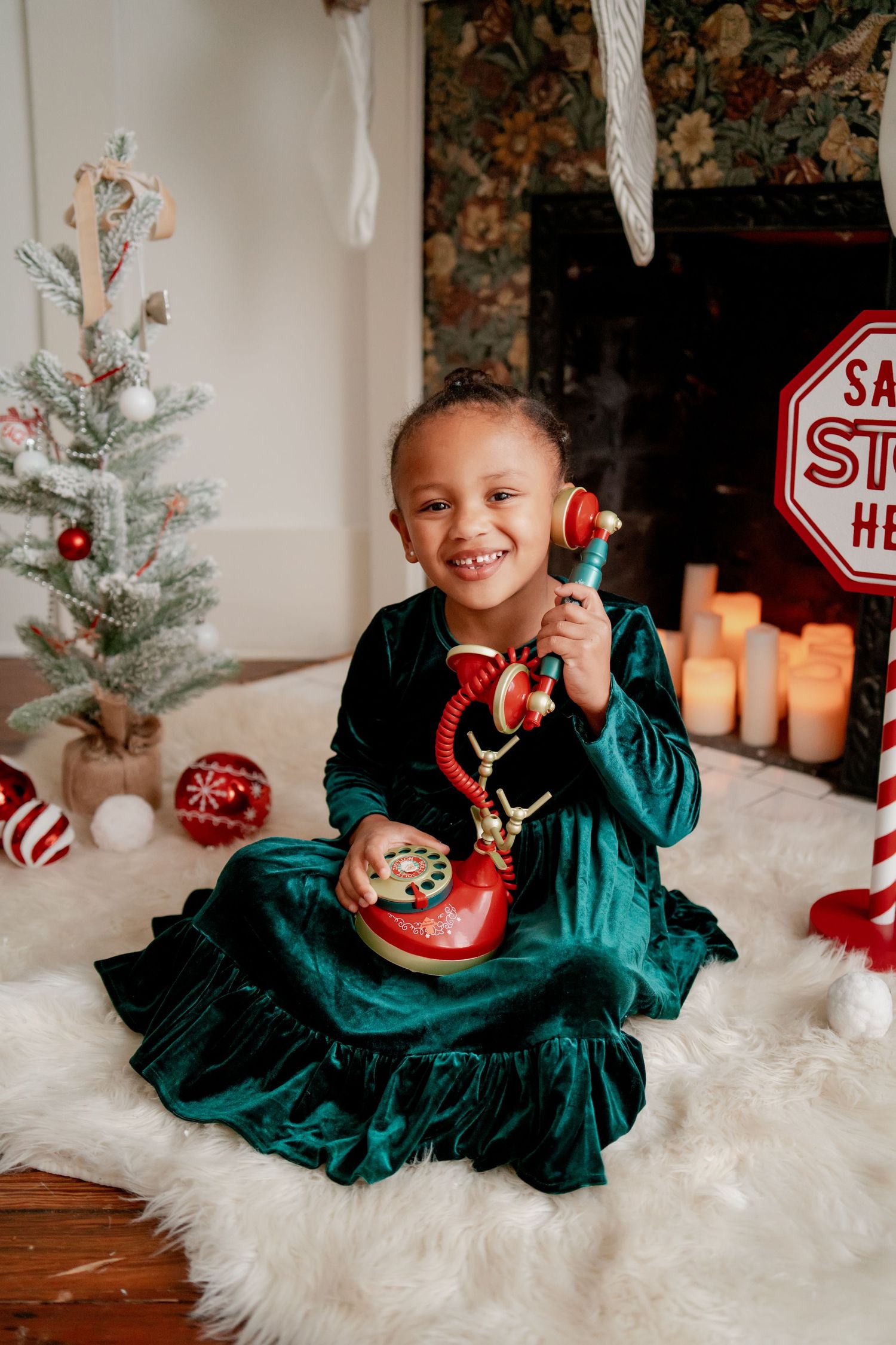 Child in green velvet dress holds vintage red phone near Christmas decorations.