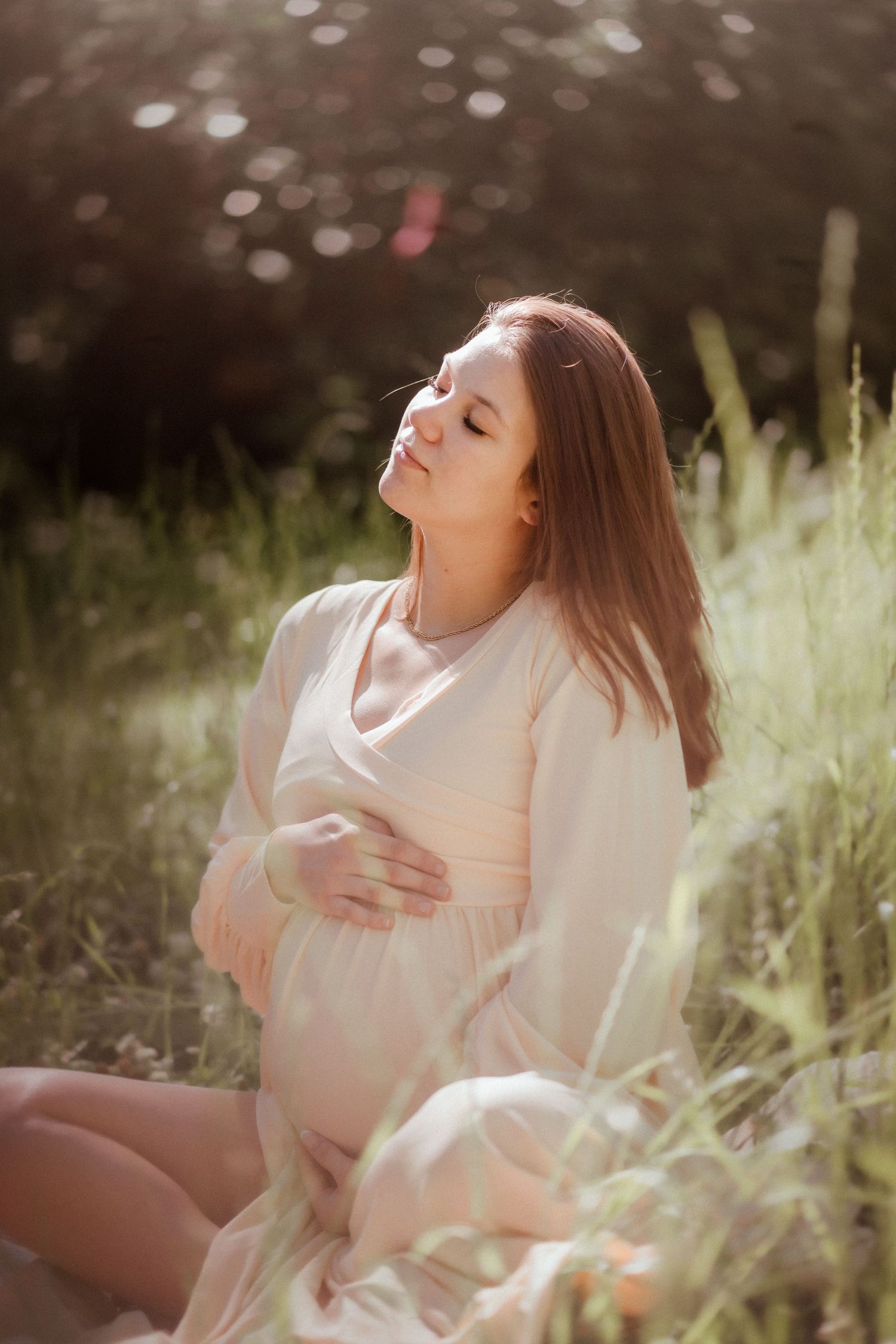 Pregnant woman in beige dress sitting in sunlit meadow, cradling her belly with eyes closed peacefully.