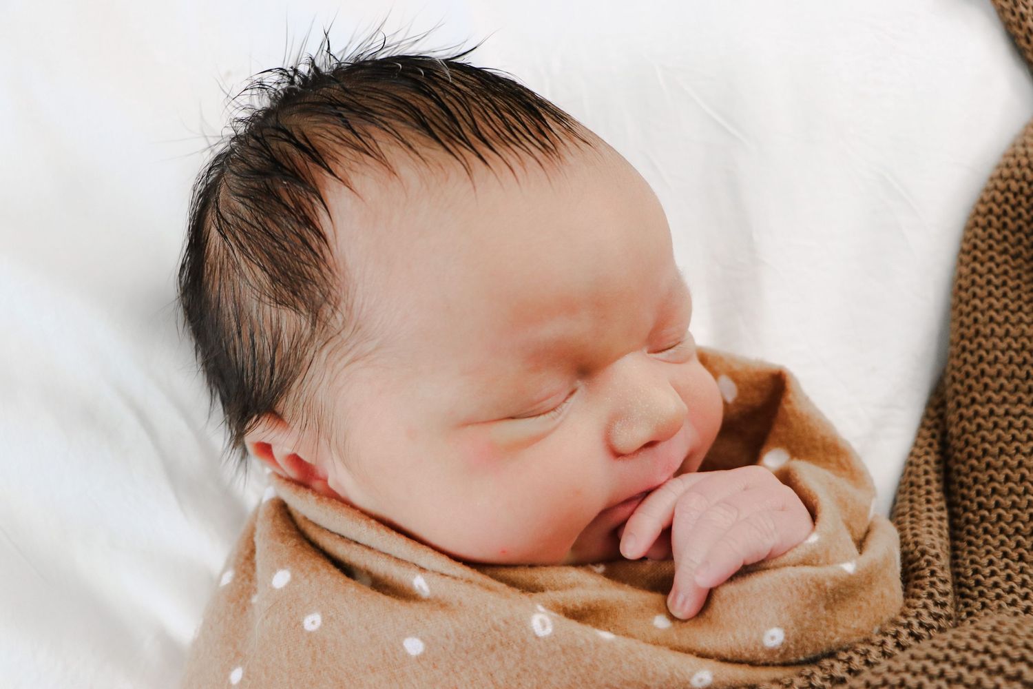 A peaceful sleeping newborn baby wrapped in white and brown blankets rests with hand near face.