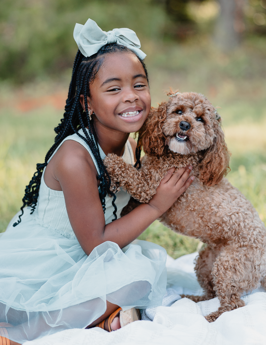 Young girl in a white dress with bow hugging brown toy poodle on blanket outdoors.