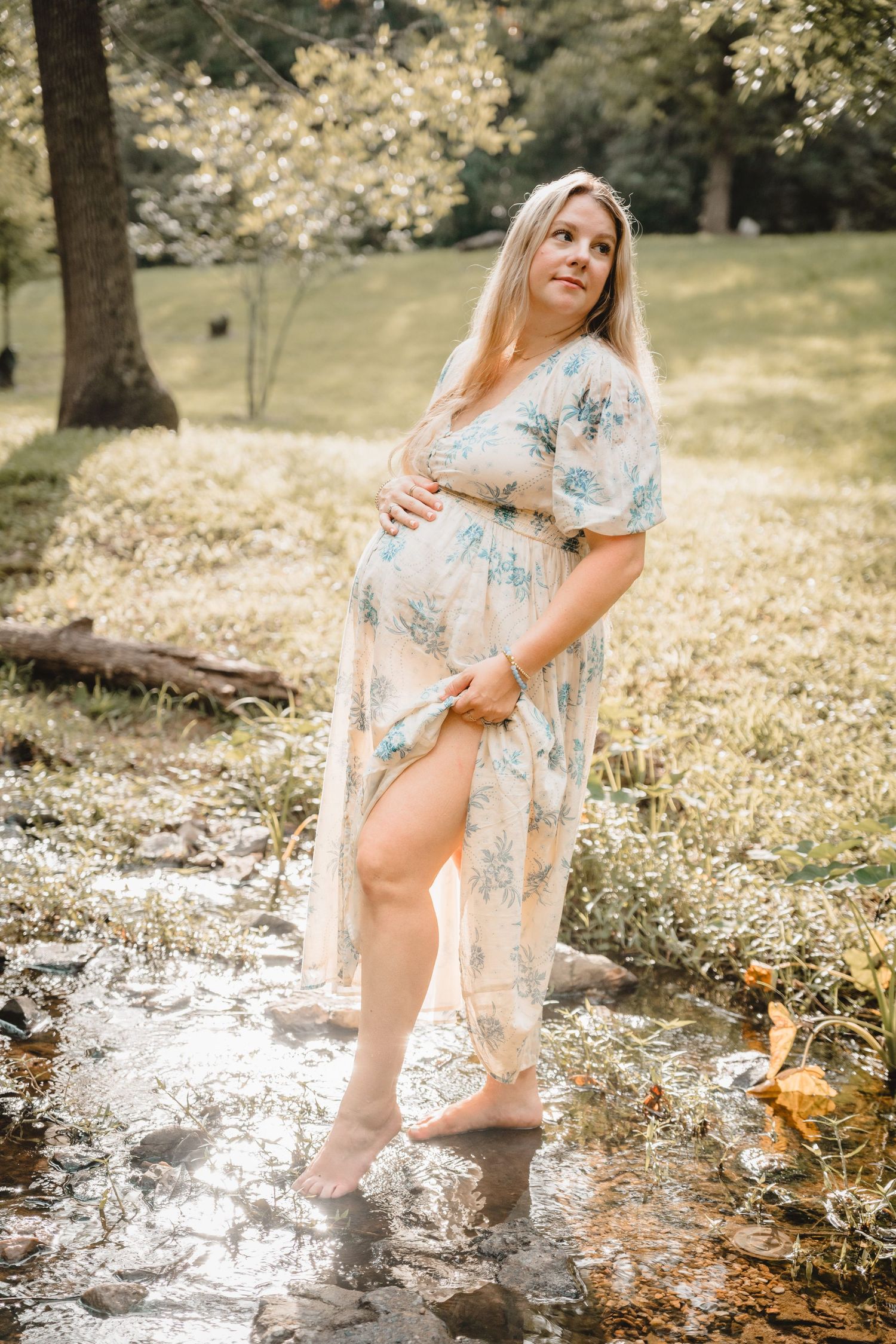 Pregnant woman in floral maxi dress standing barefoot in a creek, cradling her baby bump in a sunlit park.
