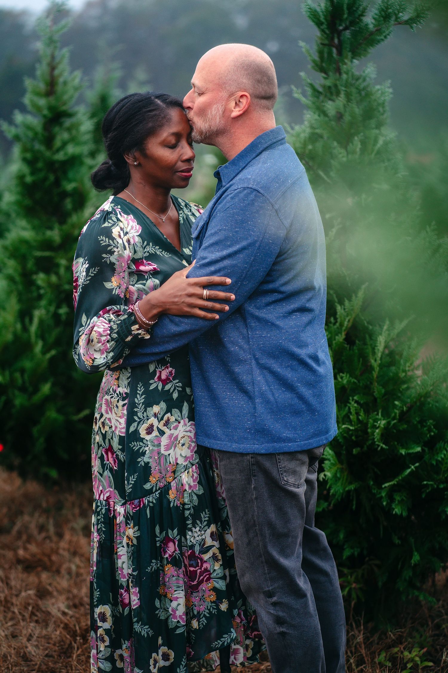 A couple embraces tenderly in front of evergreen trees during an outdoor photo session.
