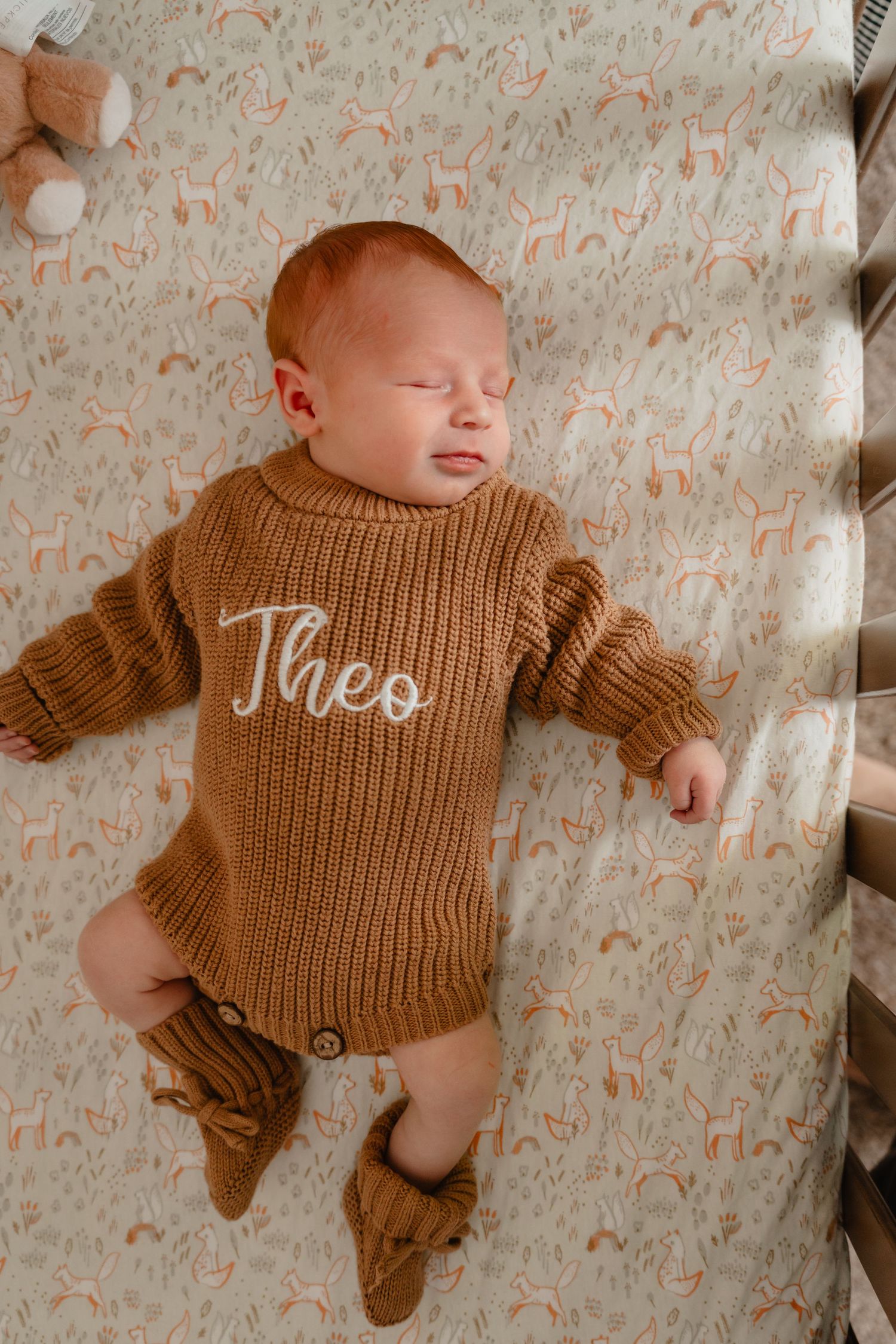 Newborn baby named Theo sleeping in a crib wearing a brown knit sweater romper with booties.