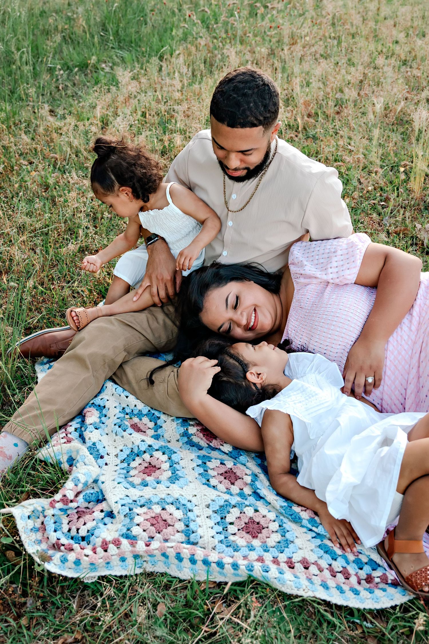 A family shares tender moments while lying on a blue and white patterned blanket in a grassy field.