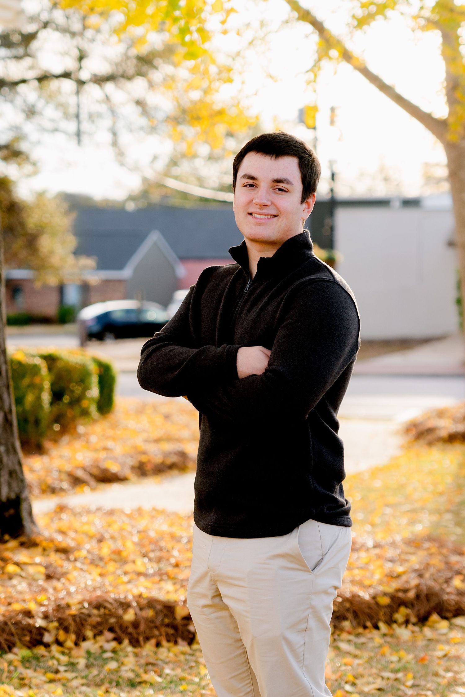A person in a black fleece jacket and khaki pants stands with crossed arms on an autumn day with yellow leaves in the background.