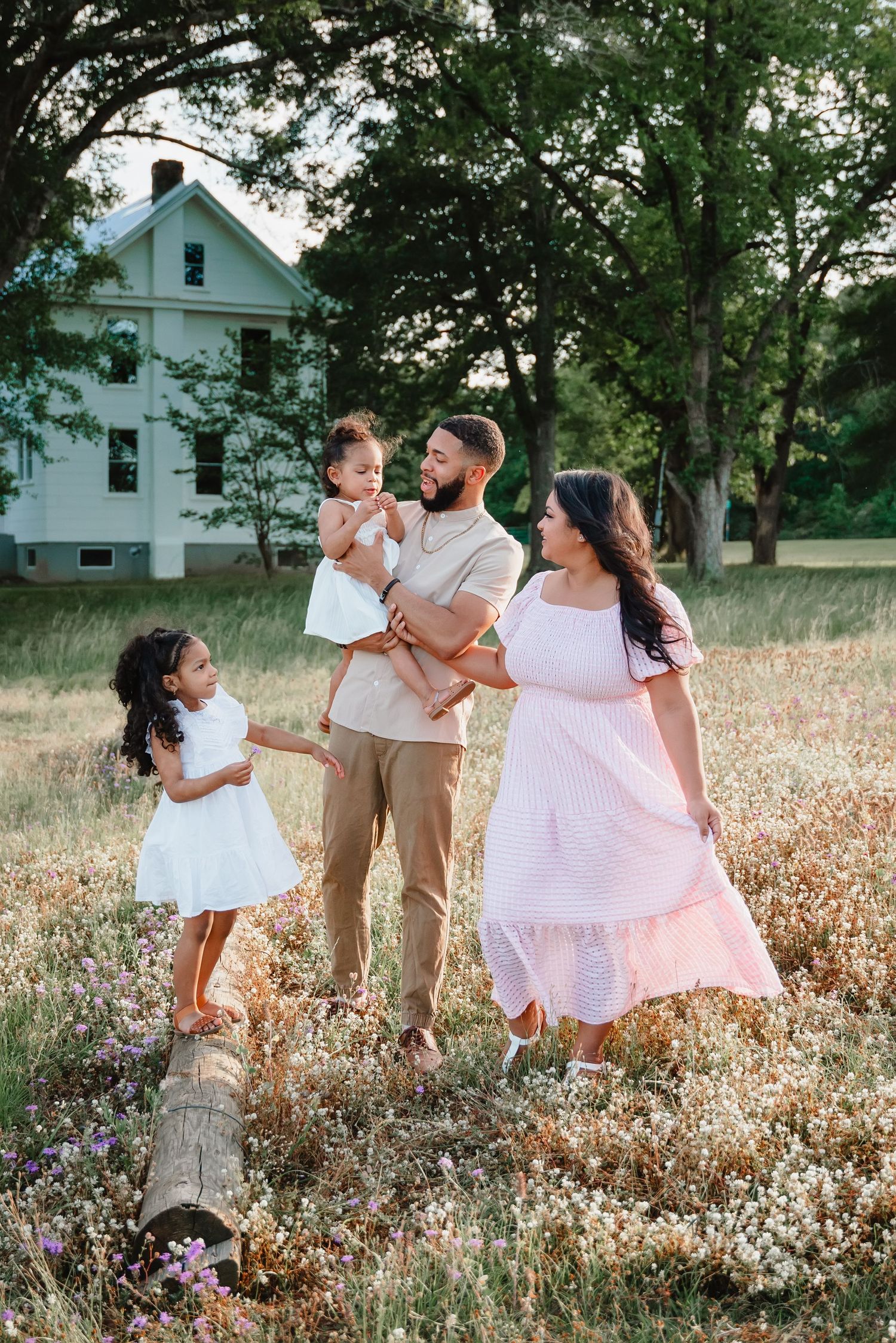 Family of four posing in a wildflower meadow near a white farmhouse, wearing coordinated neutral and white outfits.