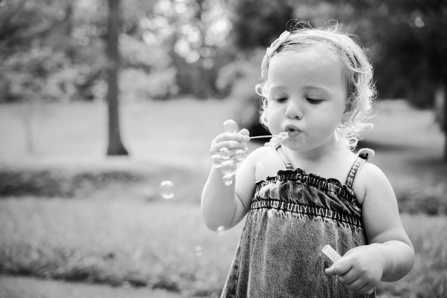 A young toddler girl blowing bubbles outdoors in a black and white photo, wearing a dress in a park setting.