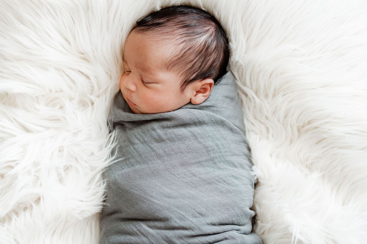 Baby swaddled in gray blanket resting peacefully on white furry blanket.