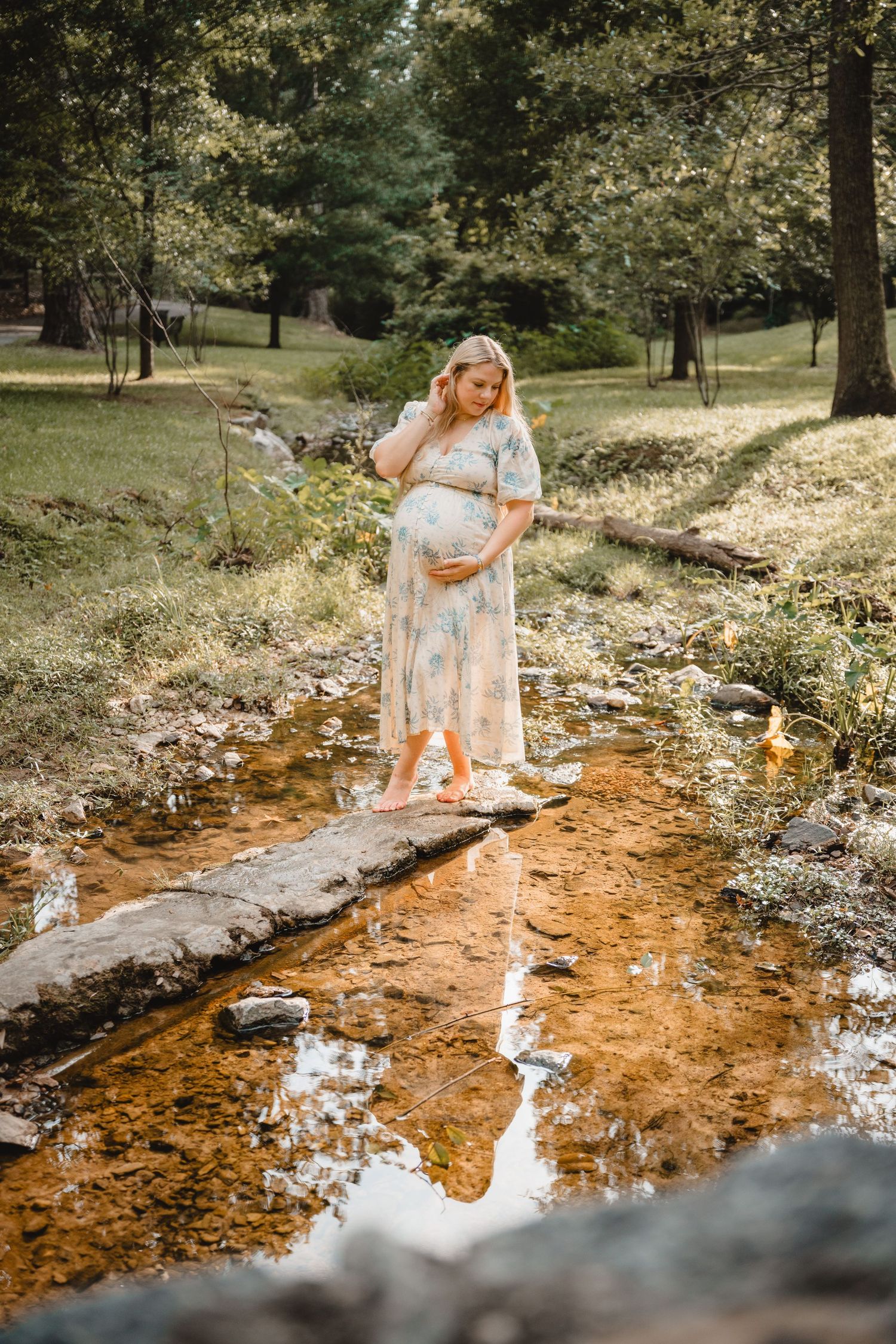 Pregnant woman in floral dress standing on stepping stones over a creek in a lush green park.
