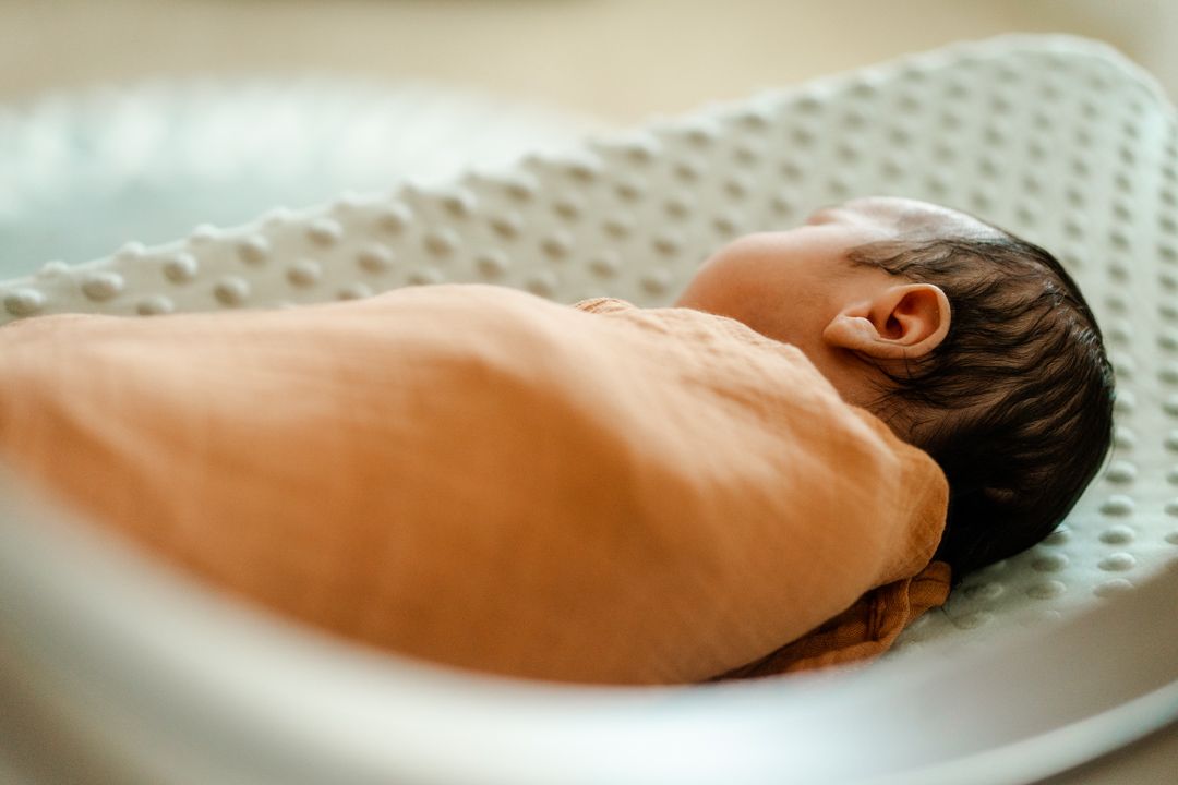 A newborn baby rests peacefully on a textured white changing pad or mattress.
