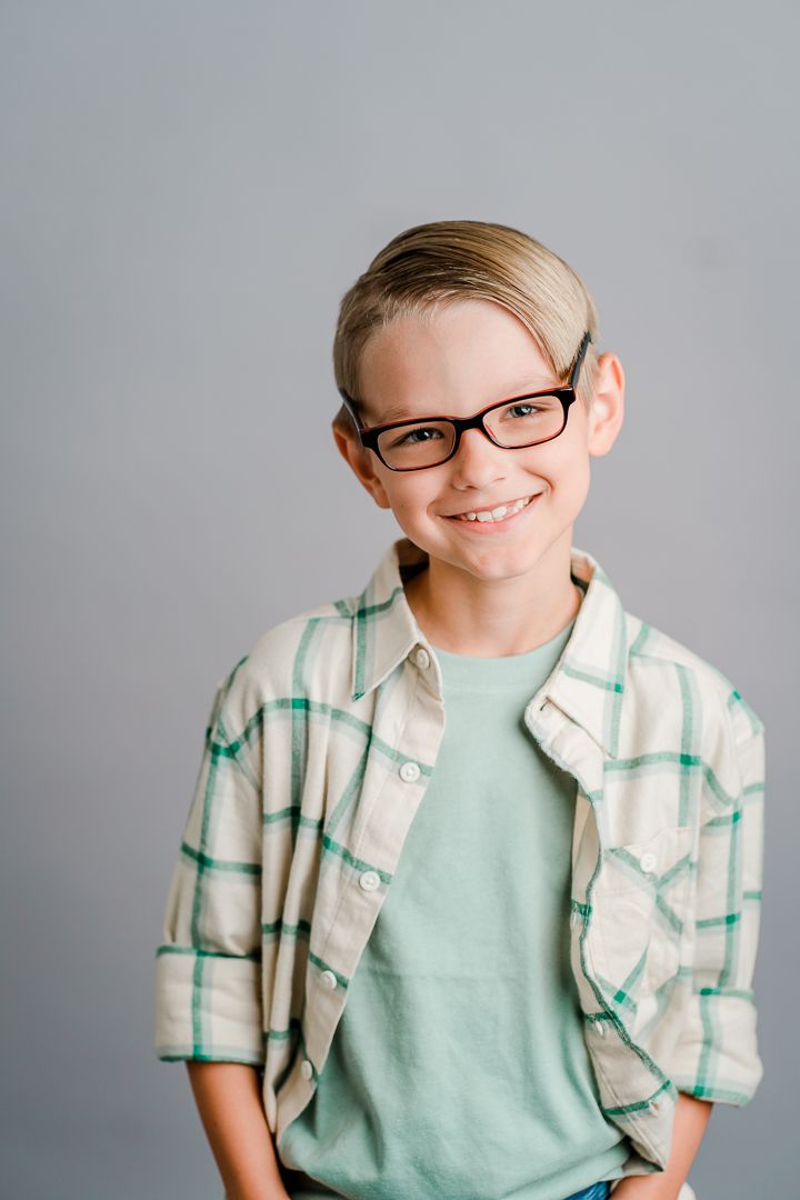 A person wearing glasses and a mint green t-shirt with a plaid button-up shirt smiles against a gray background.