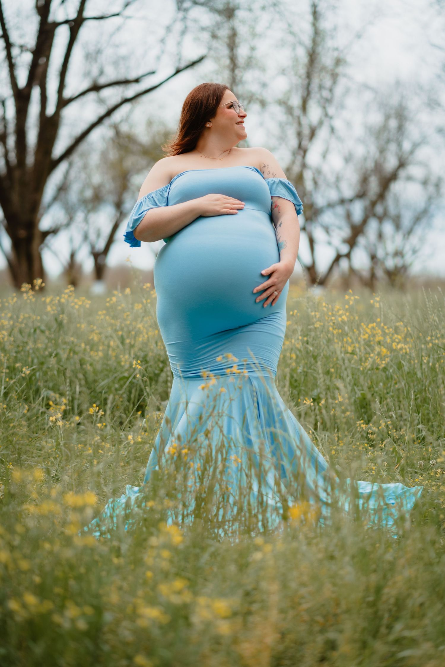 A pregnant woman stands in a field of yellow flowers, wearing a flowing blue dress and smiling at the camera.