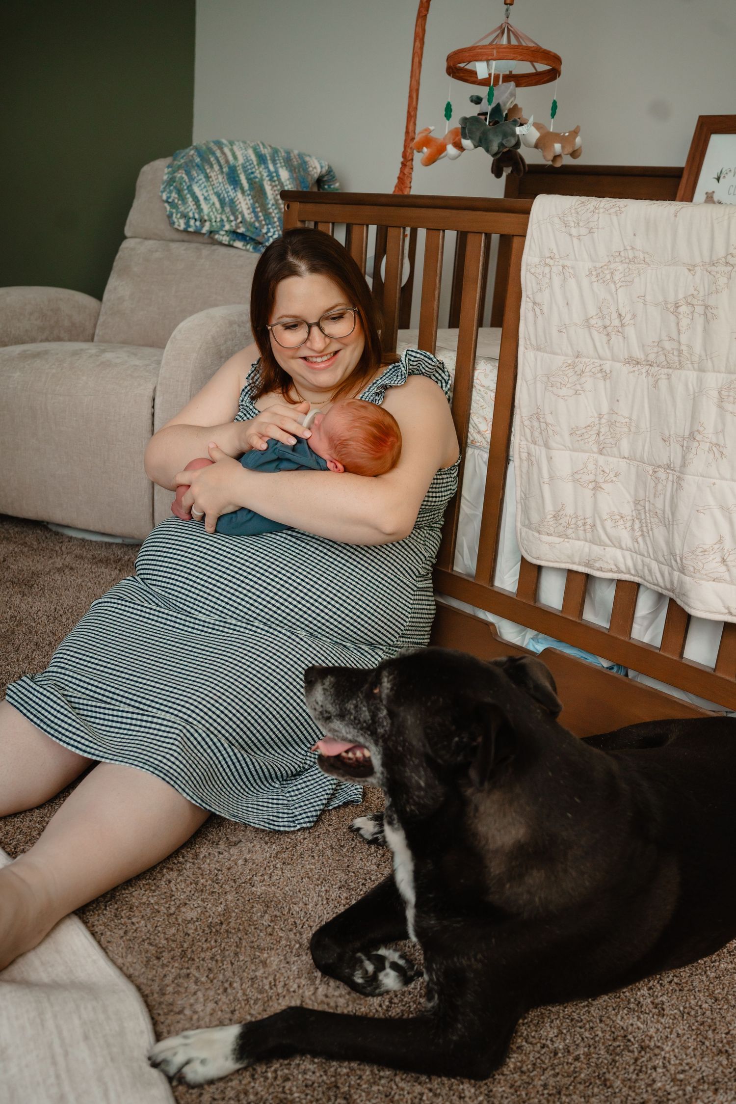 A smiling mother holds her newborn baby while sitting on the floor next to a black dog and wooden crib during in-home session Kathleen GA.