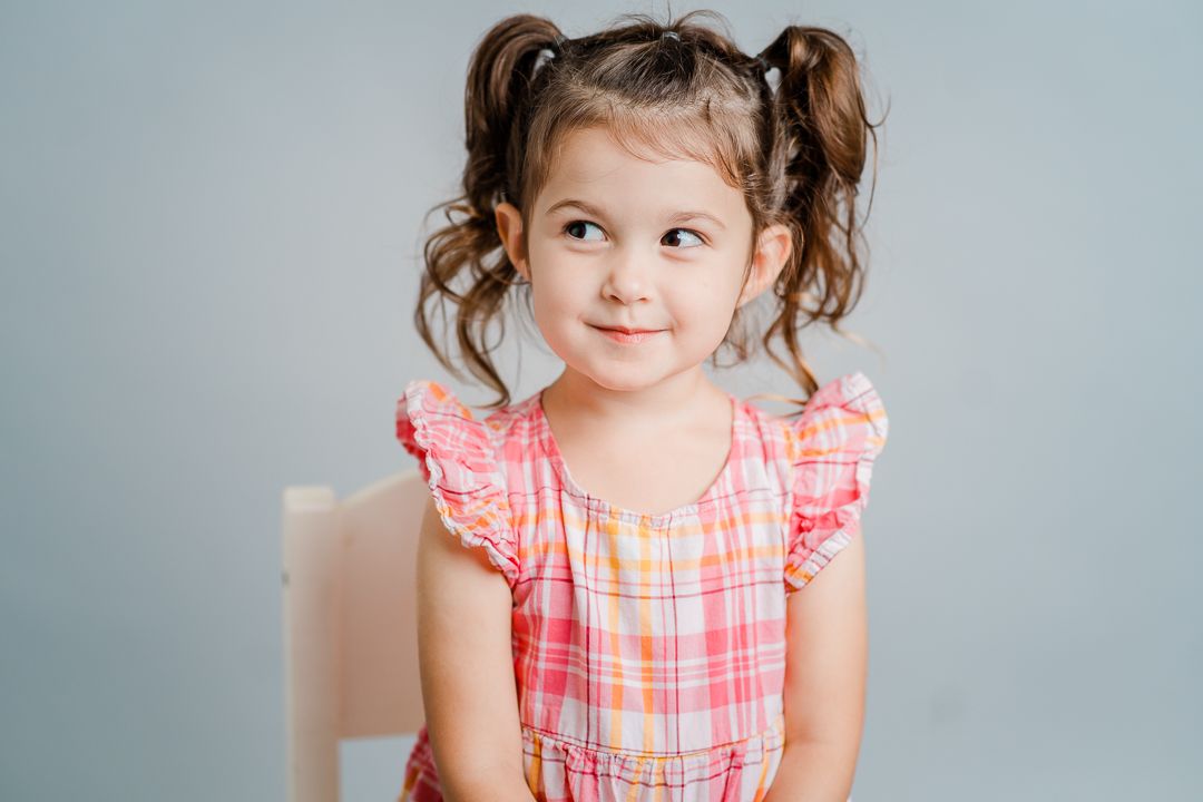 A young child in a pink plaid dress makes various playful expressions while posing against a light gray background.